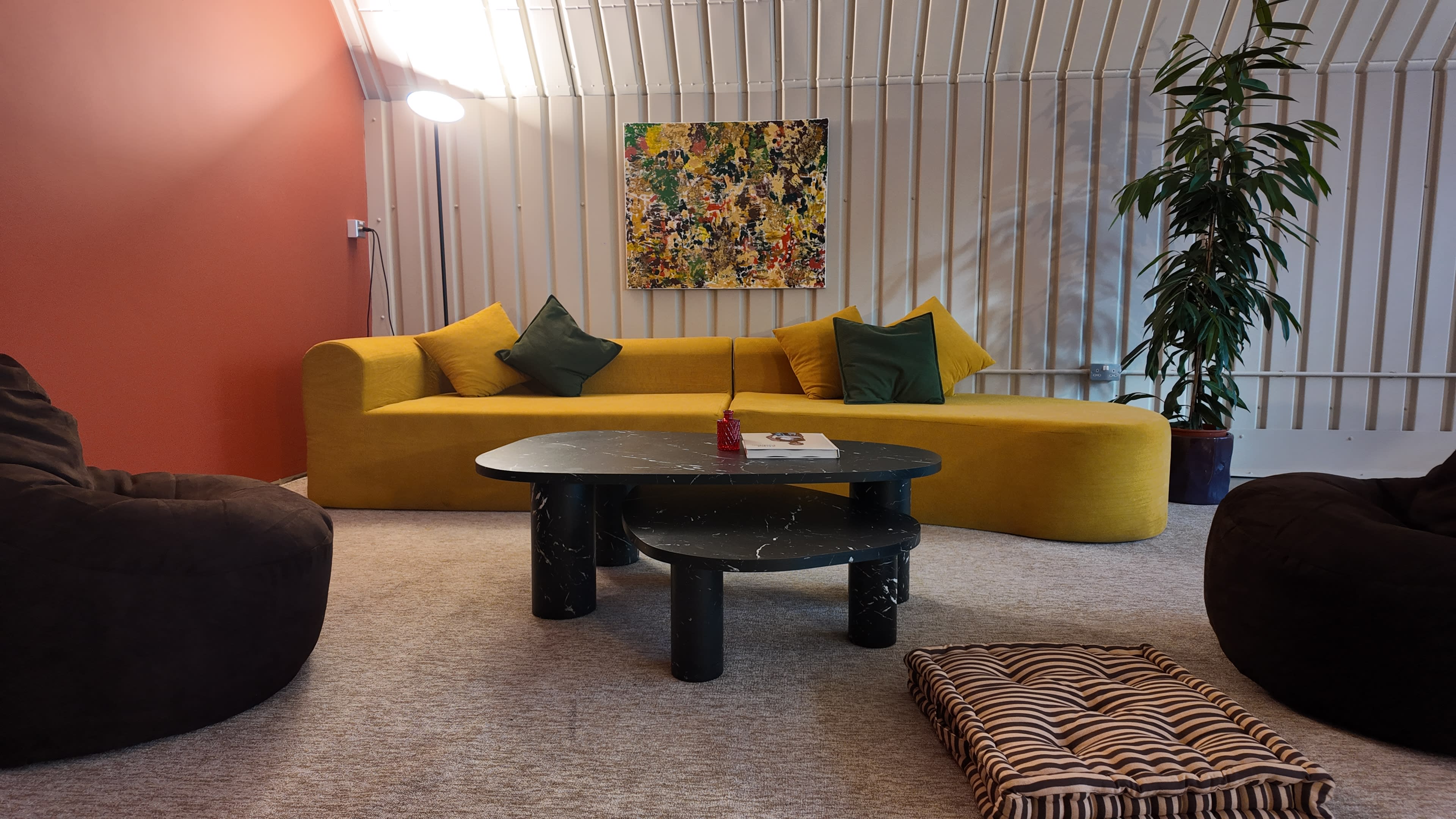 The image shows a modern living space featuring a yellow curved sofa, a black marble coffee table, and a large abstract painting on the wall, along with a plant and comfortable seating.