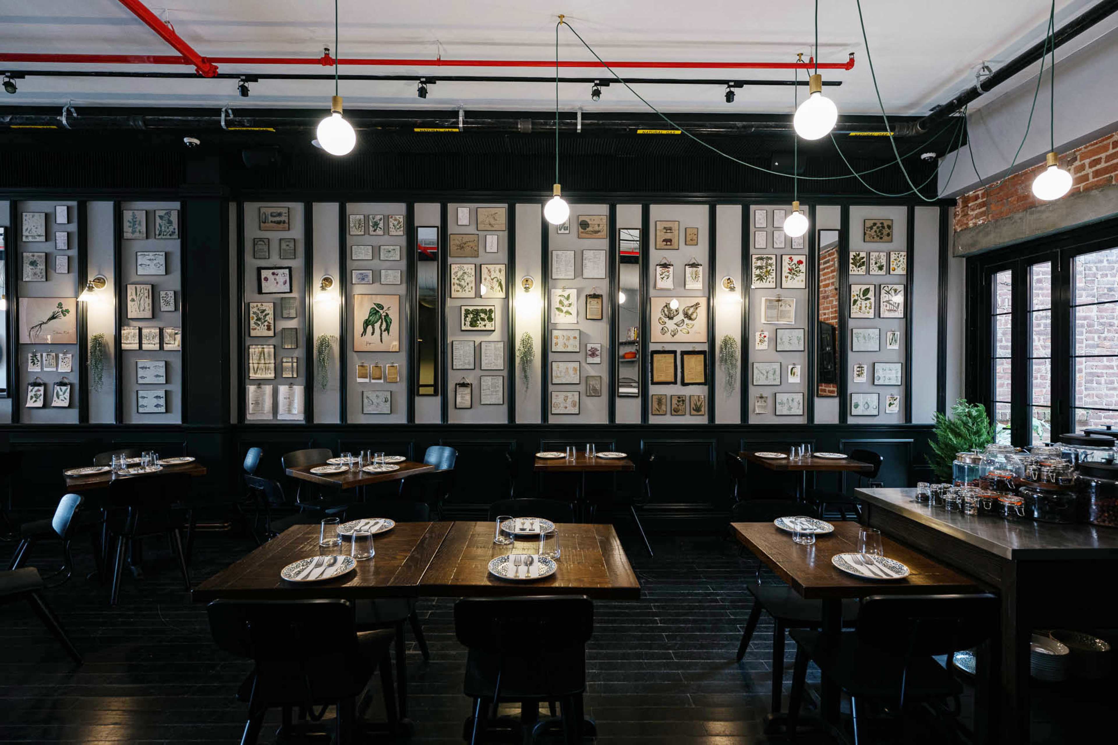 The image shows a contemporary restaurant interior featuring dark wood tables, black chairs, and a wall adorned with framed artwork and botanical illustrations.