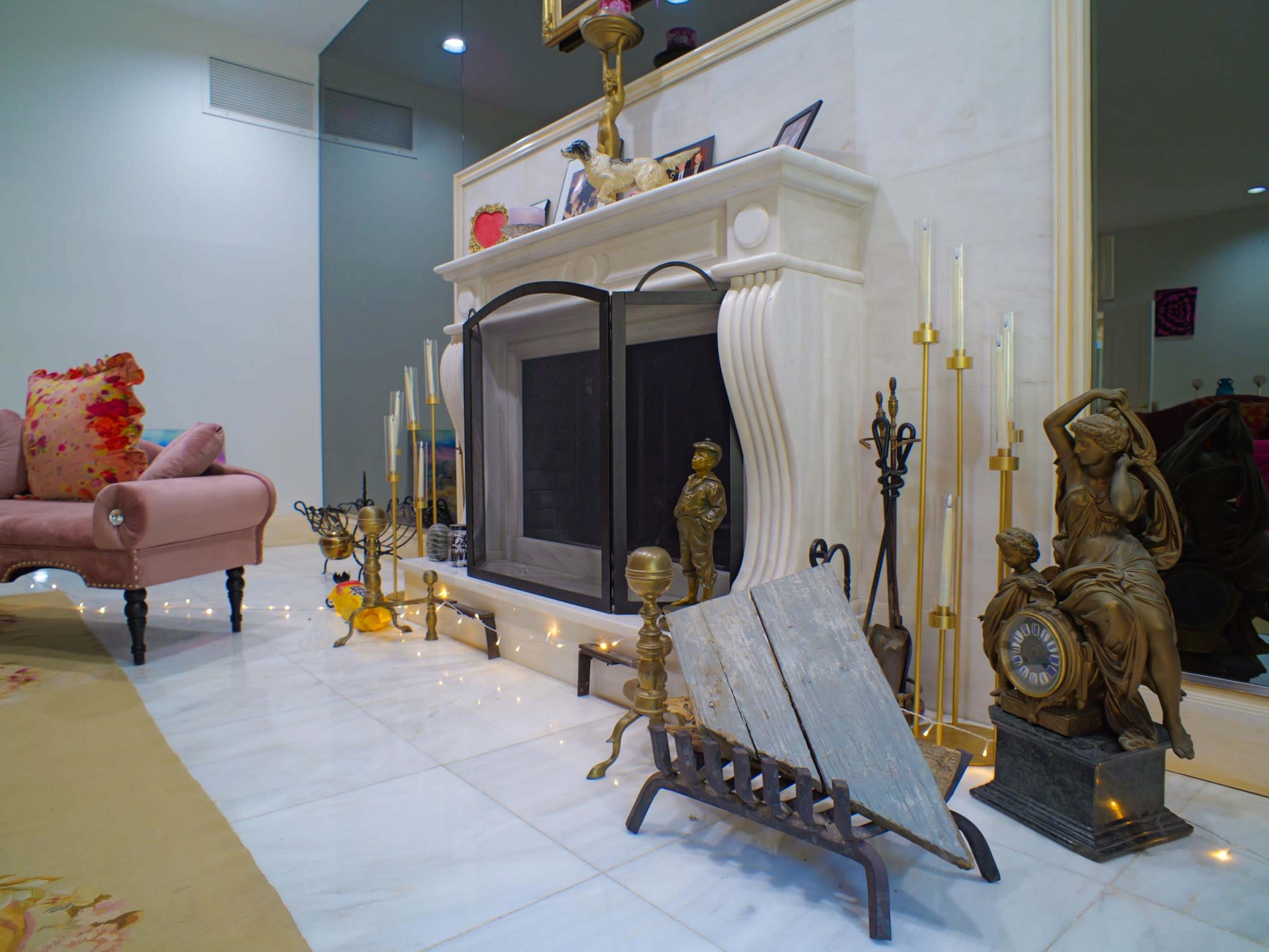 The image shows a well-decorated living room featuring a marble fireplace with various decorative items, including candlesticks and a wooden log holder, alongside a pink armchair and a plush area rug.