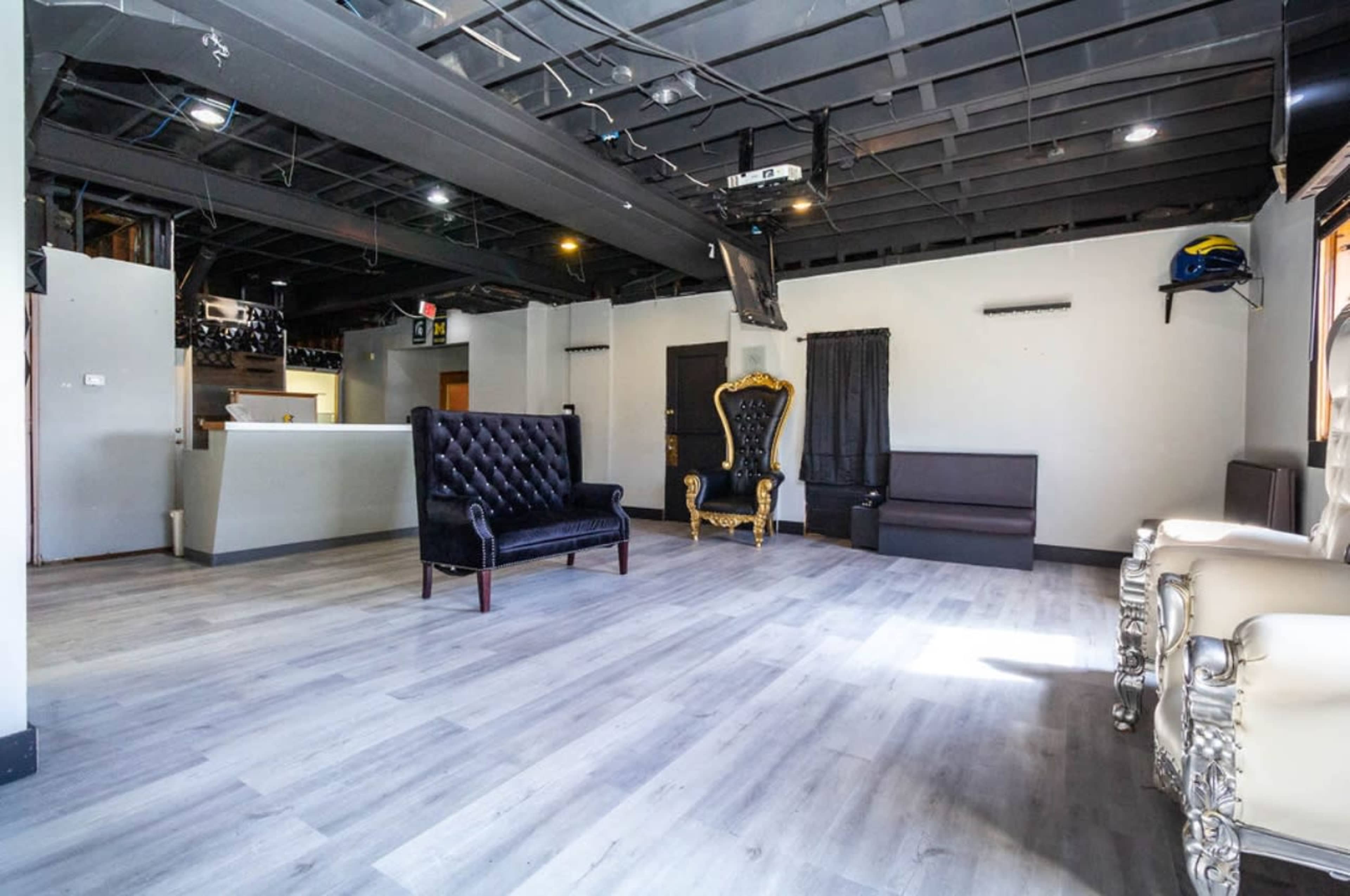 The image shows an empty interior space featuring a mix of seating options, including a black tufted sofa and a decorative golden chair, with grey laminate flooring and exposed ceiling beams.