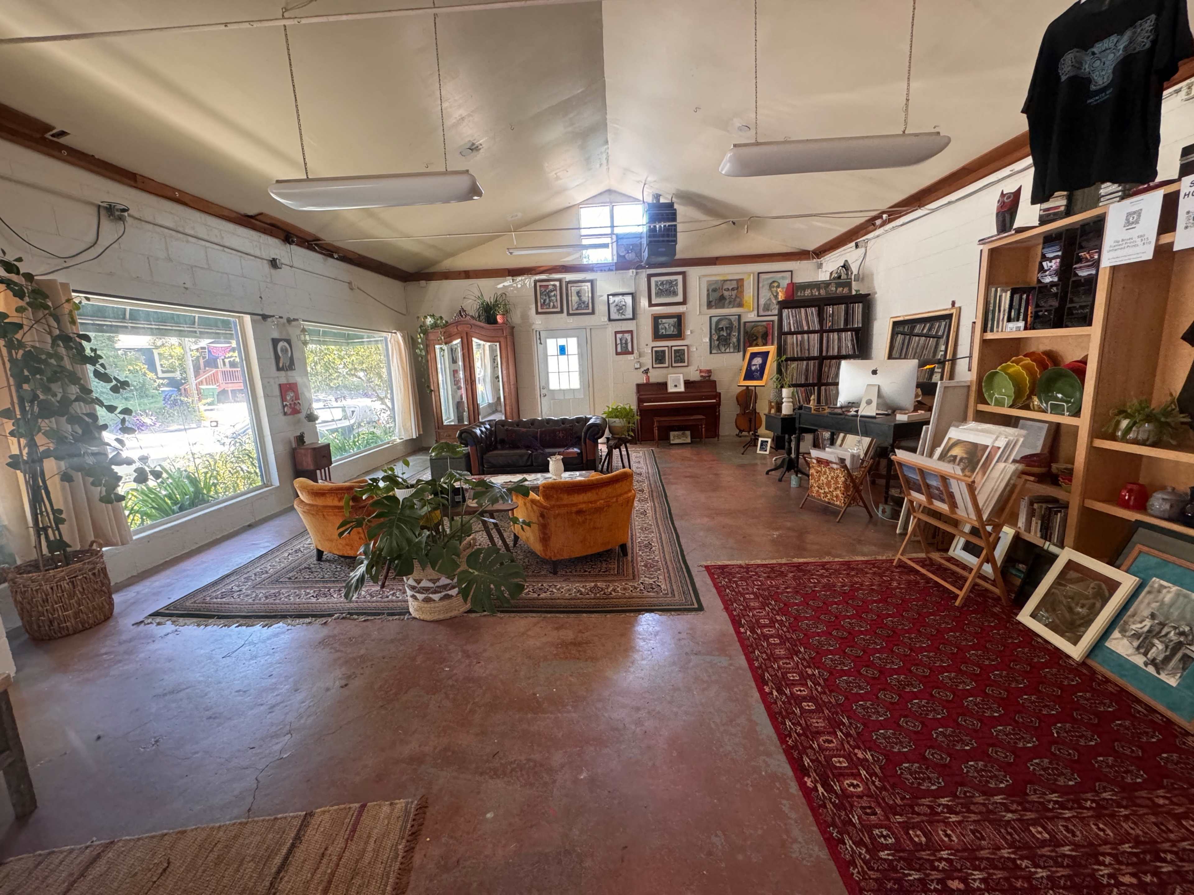 A spacious, well-lit room featuring a blend of seating areas, shelves filled with books and artworks, and various plants, all arranged around a central rug.