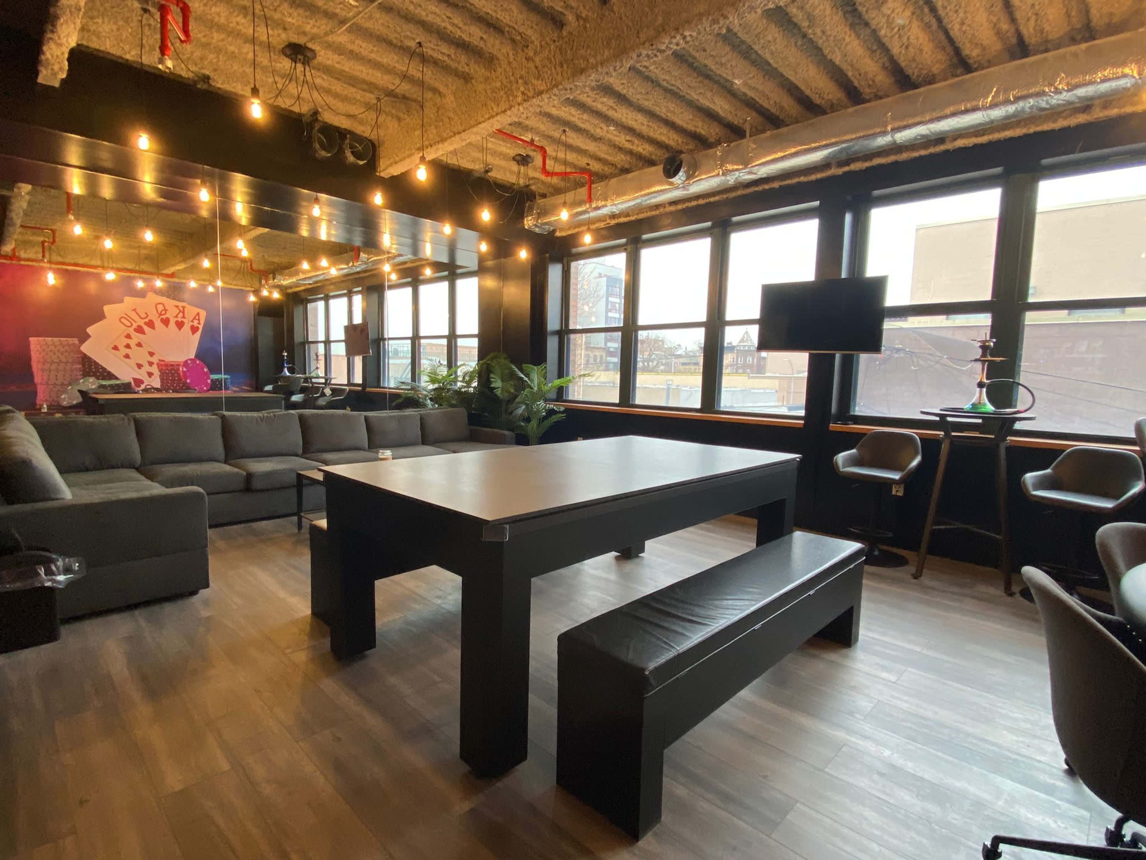 A modern office space featuring a large black table with a bench, surrounded by comfortable seating and large windows allowing natural light to fill the room.