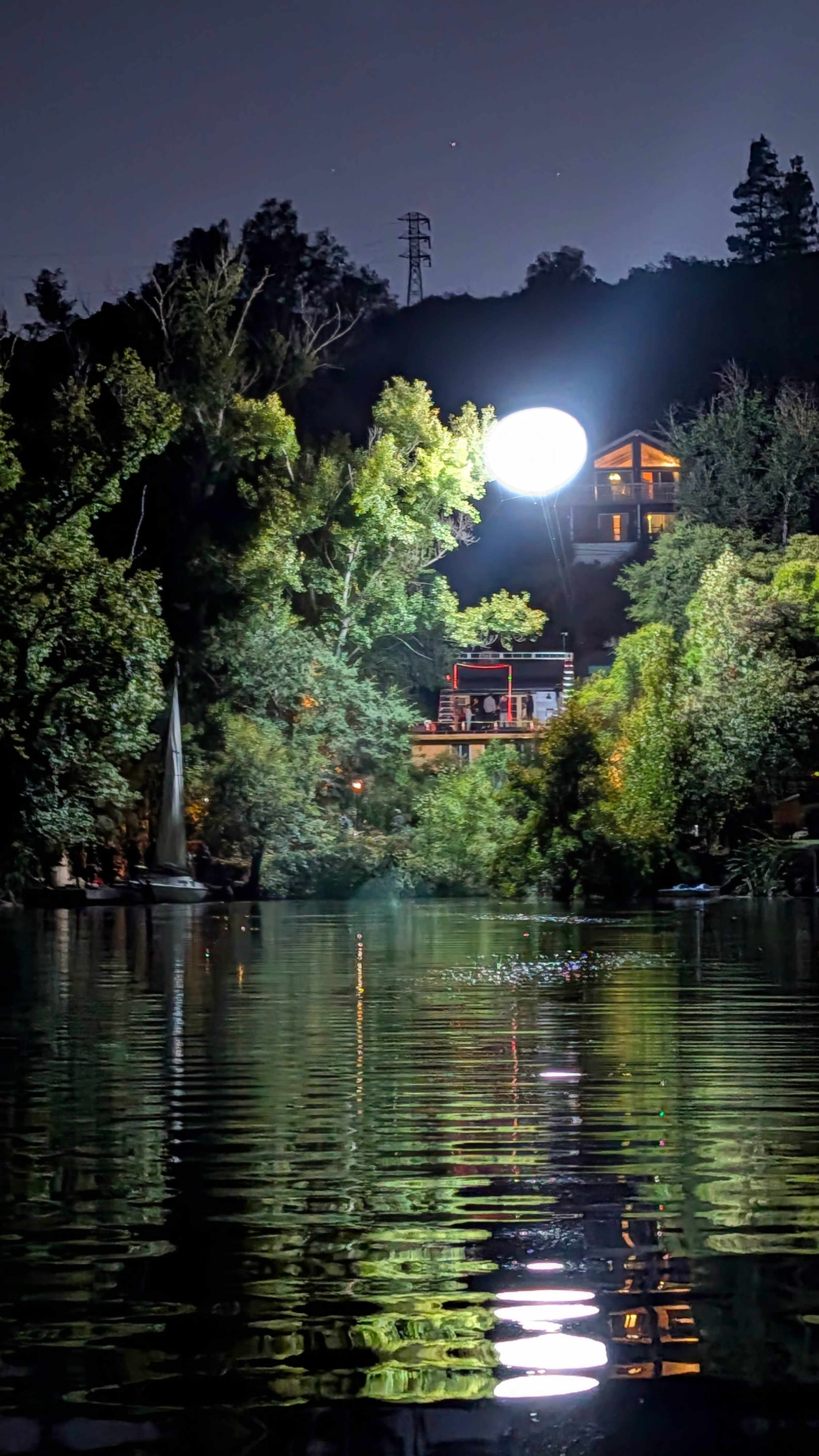 A large light is illuminating a hillside with trees and a house overlooking a calm river at night.