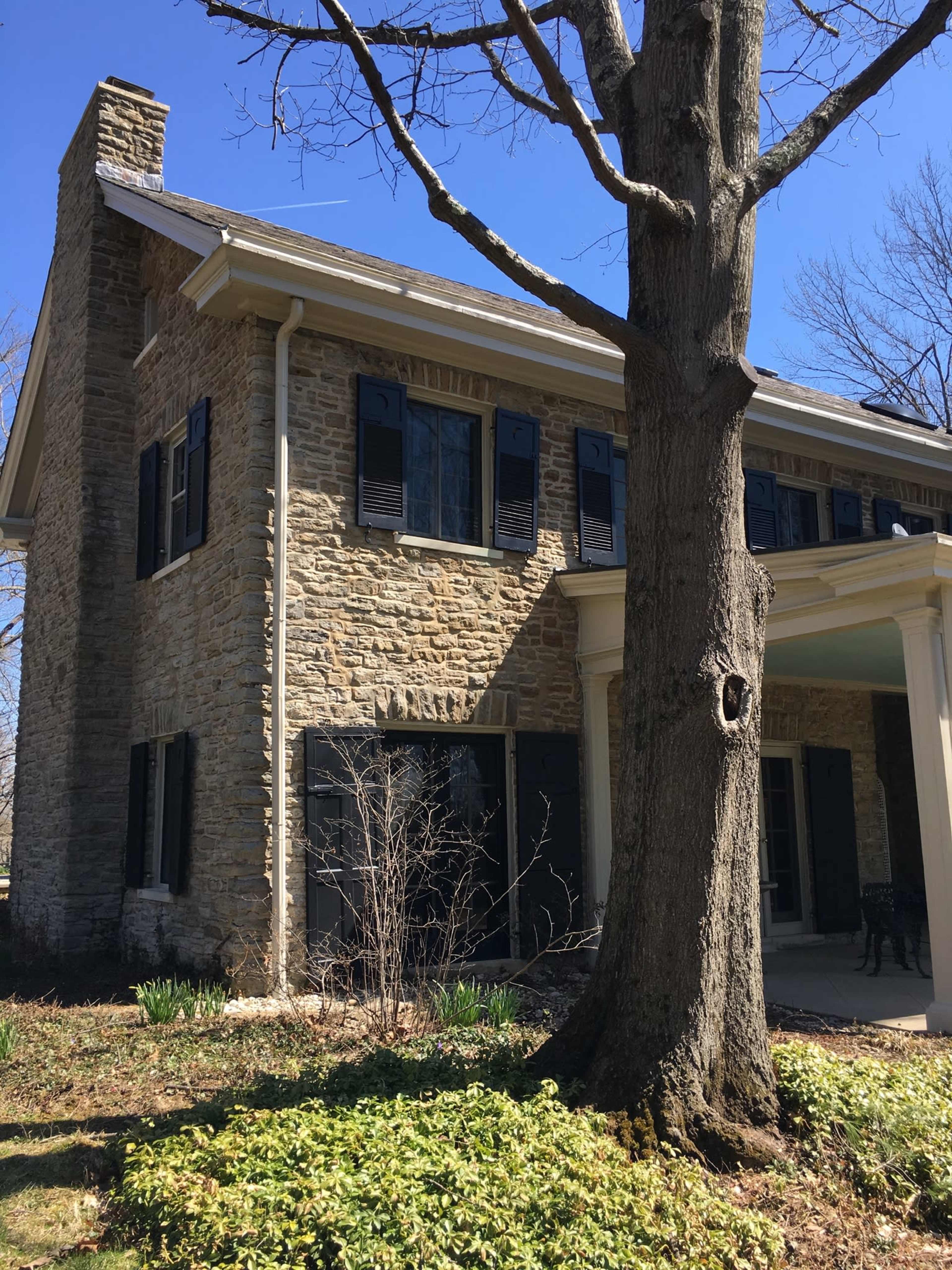 A stone house with dark shutters stands beside a large tree and is surrounded by a grassy area.