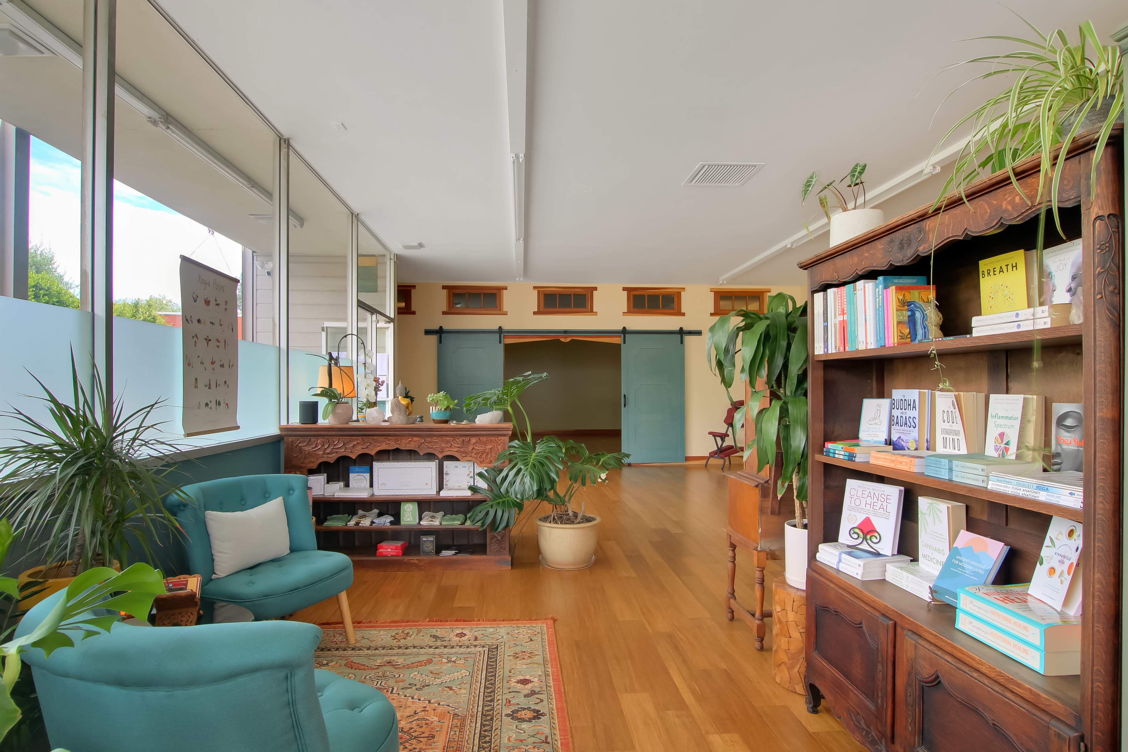 The image shows a cozy room with a wooden bookshelf filled with books, green plants, and comfortable seating, along with a decorative rug on the floor.