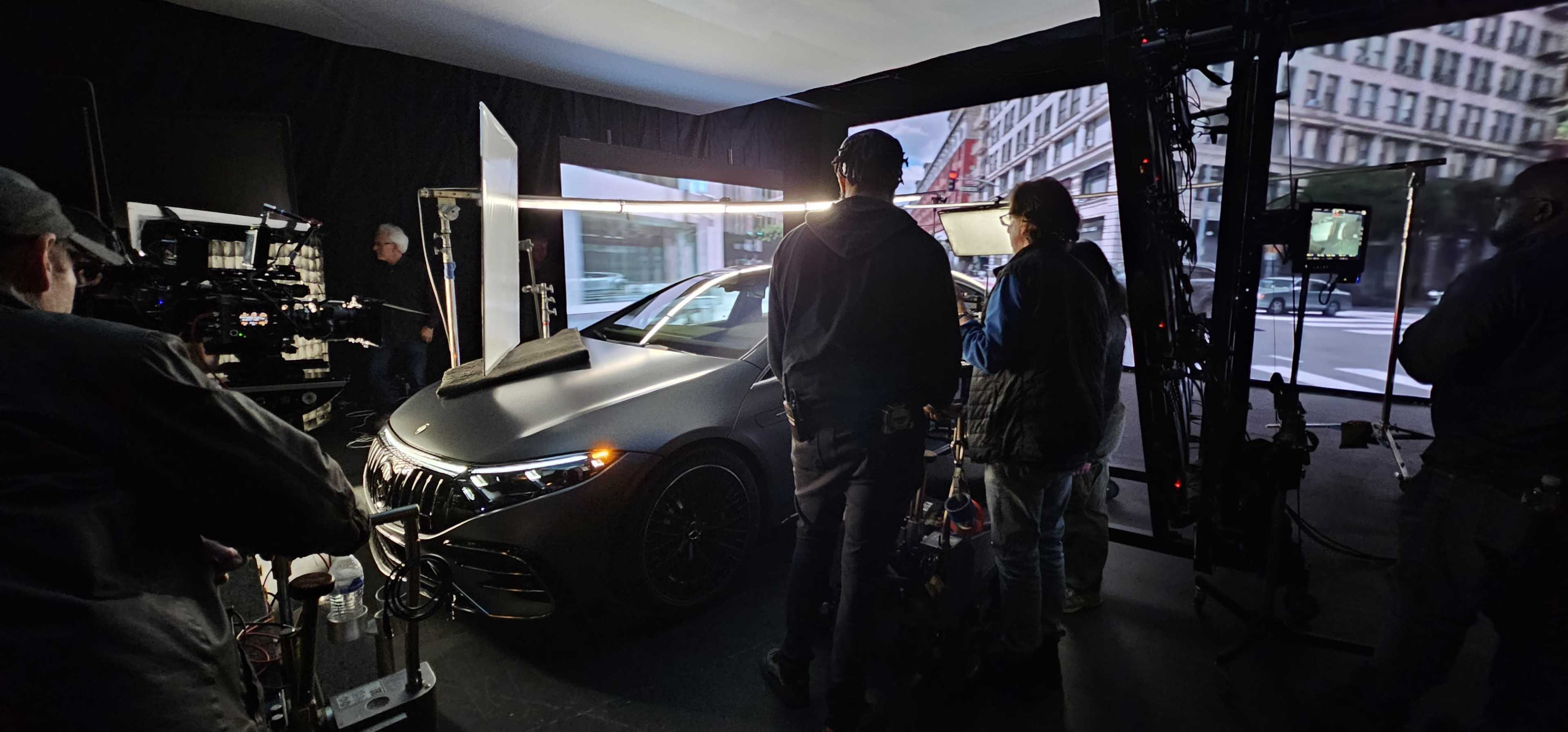 A film crew is set up inside a studio, preparing to shoot a scene featuring a car under controlled lighting conditions.