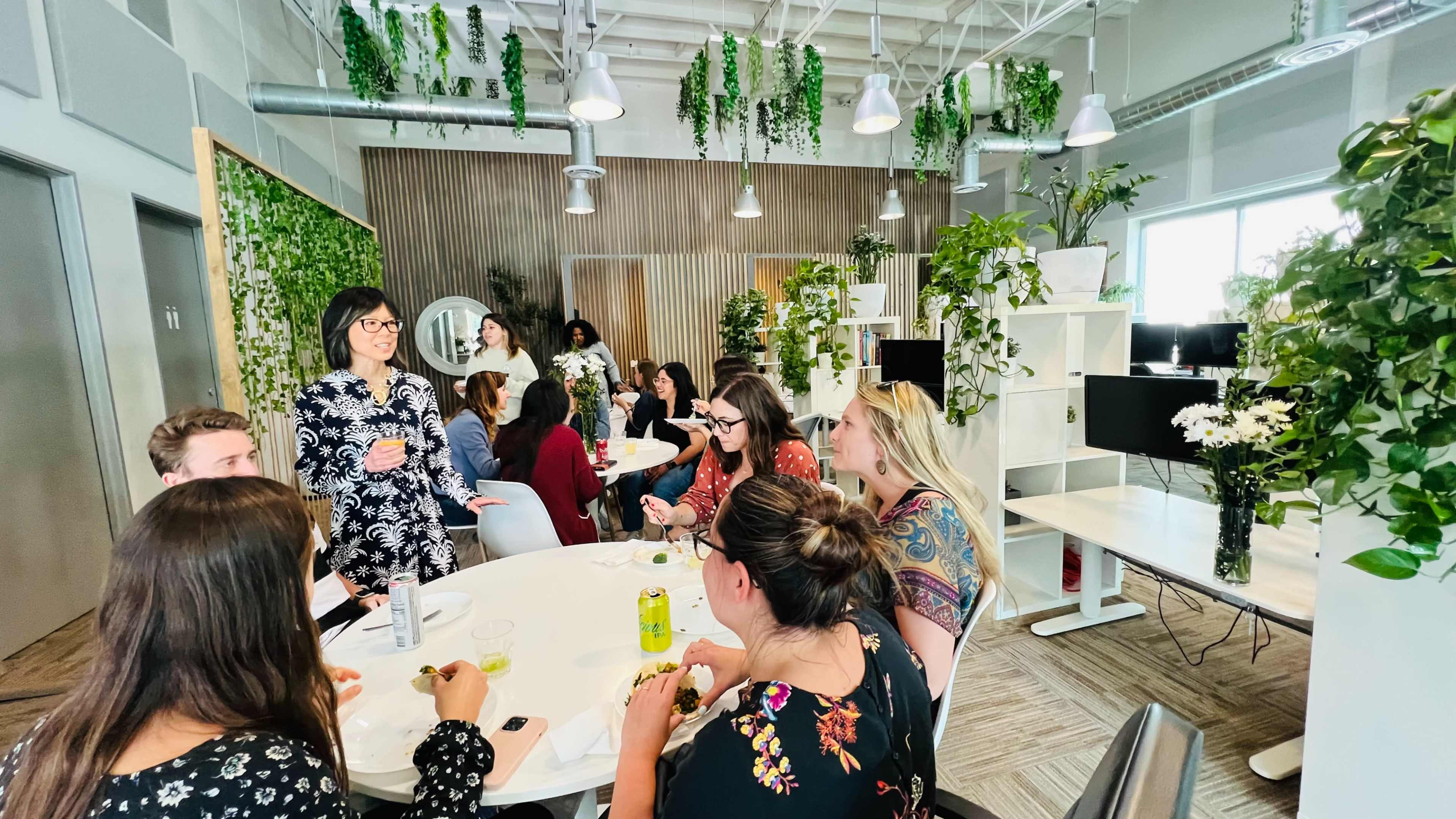 A group of people gathers around a table in a bright, plant-decorated office space, sharing food and conversation.