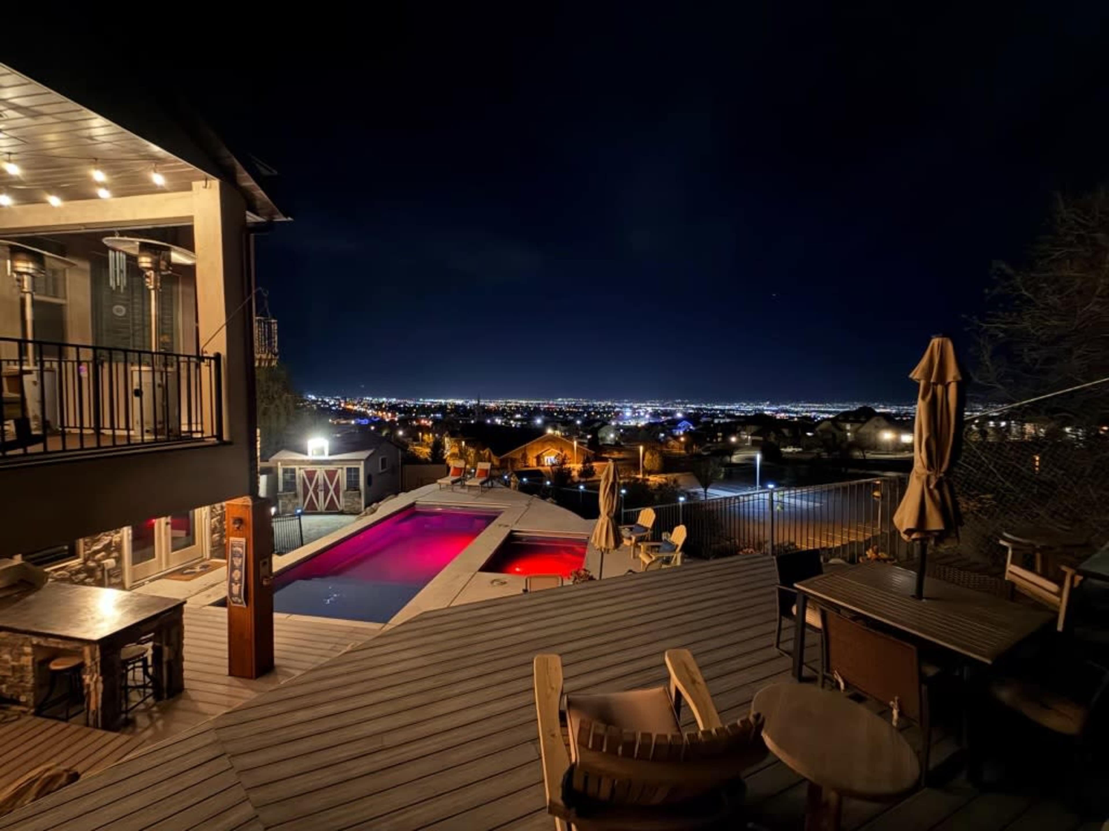 The image shows a modern outdoor patio area with a swimming pool illuminated in red, overlooking a city skyline at night.