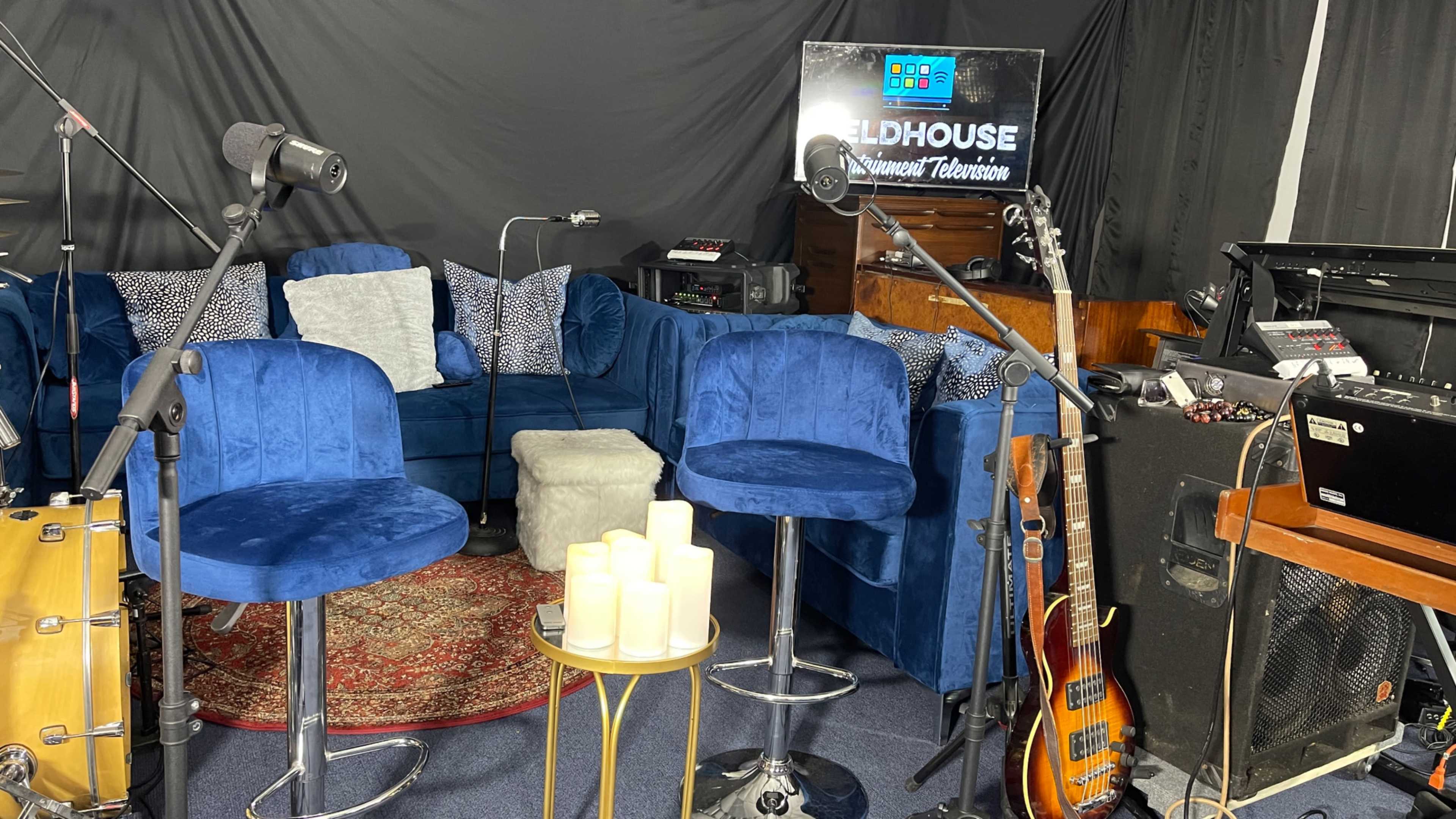 The scene shows a cozy studio setup featuring blue chairs, a small table with candles, musical instruments, and a television screen displaying the "Fieldhouse" logo.