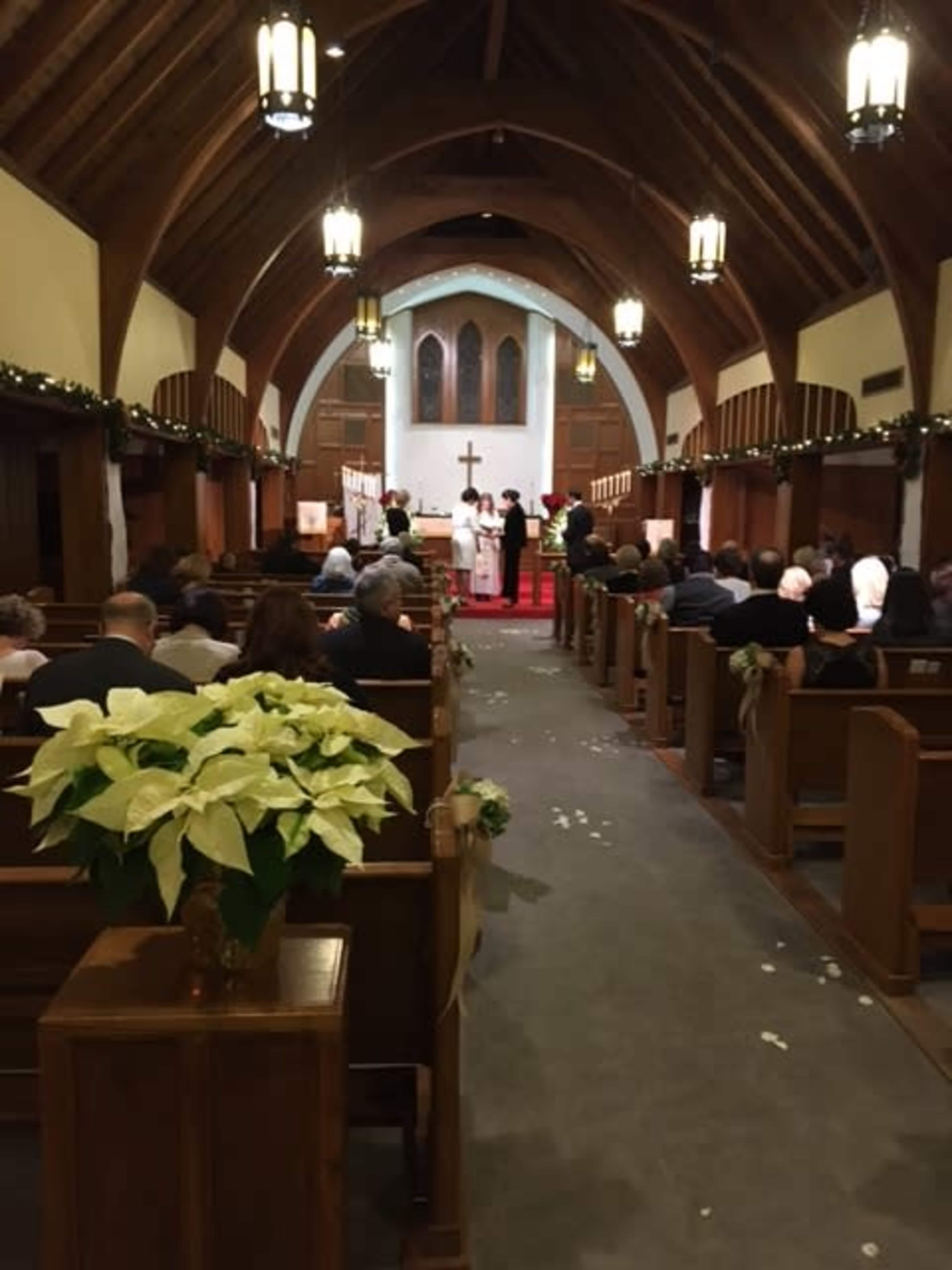 A wedding ceremony takes place inside a church, with guests seated in wooden pews and decorations featuring greenery and white flowers.