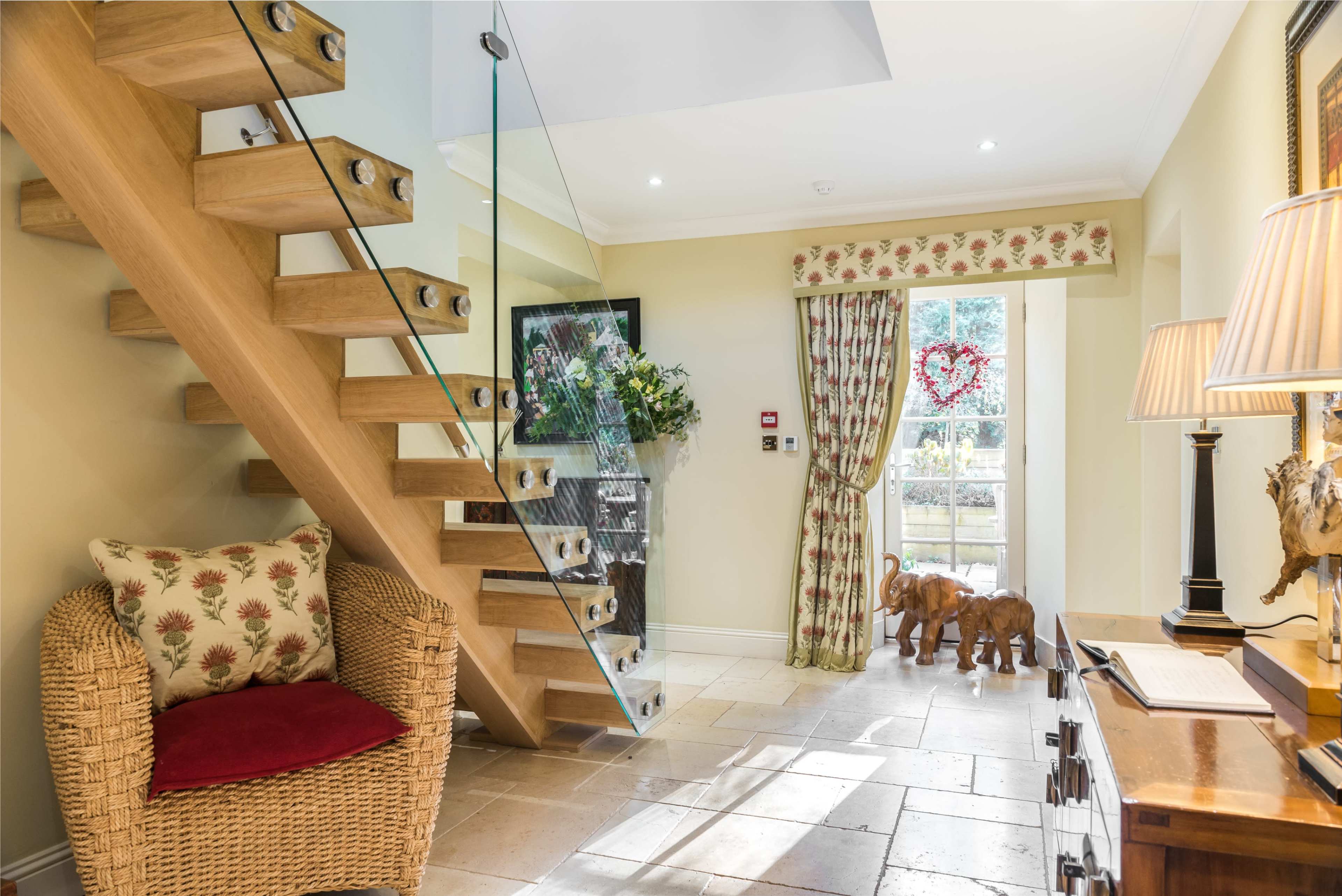An entrance hallway with a glass-paneled staircase, a wicker chair, decorative curtains, and two wooden elephant figurines near a doorway leading outside.