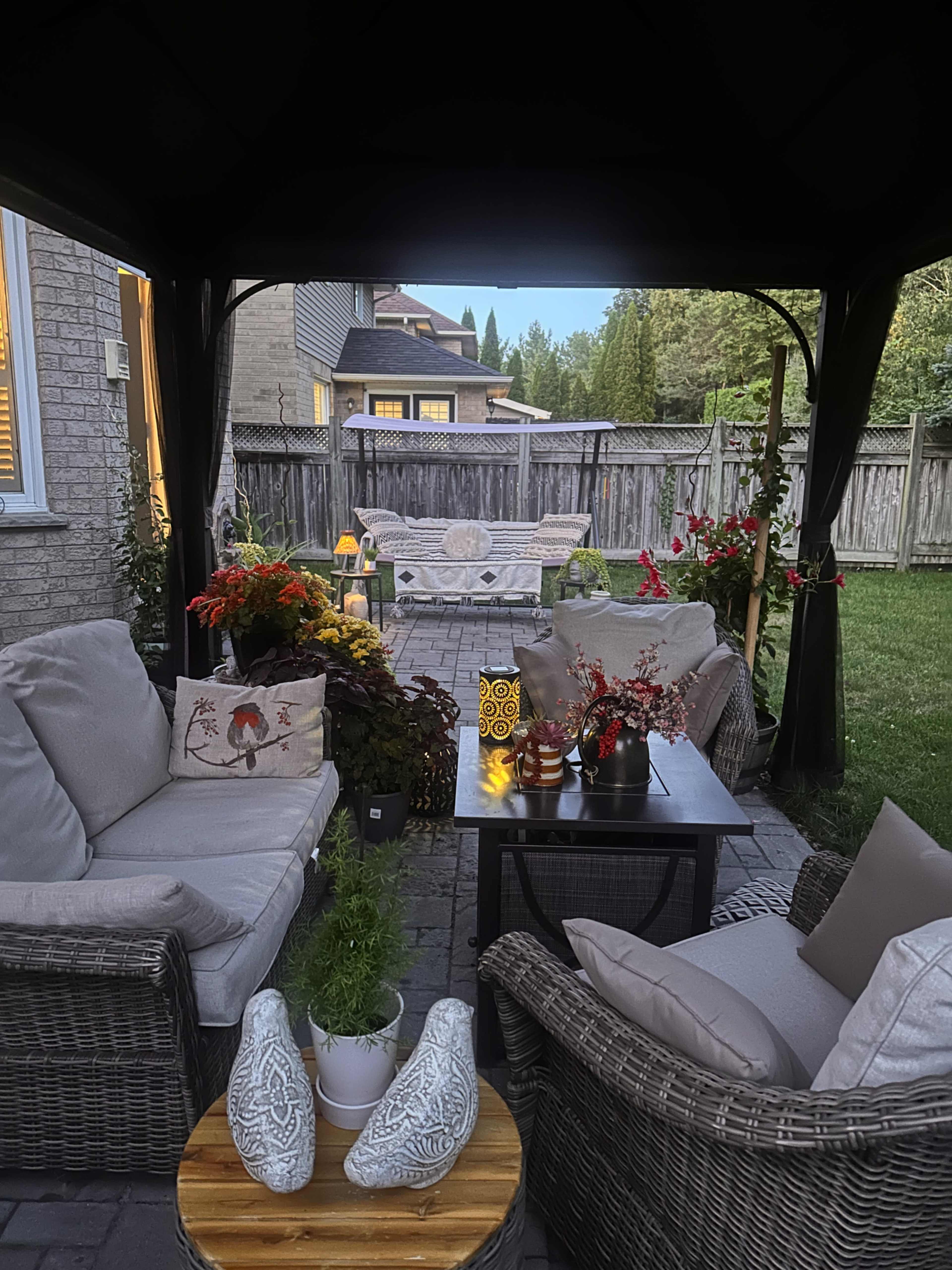 The image shows a cozy outdoor seating area with woven furniture arranged around a small table, surrounded by potted plants and colorful flowers in a backyard setting.