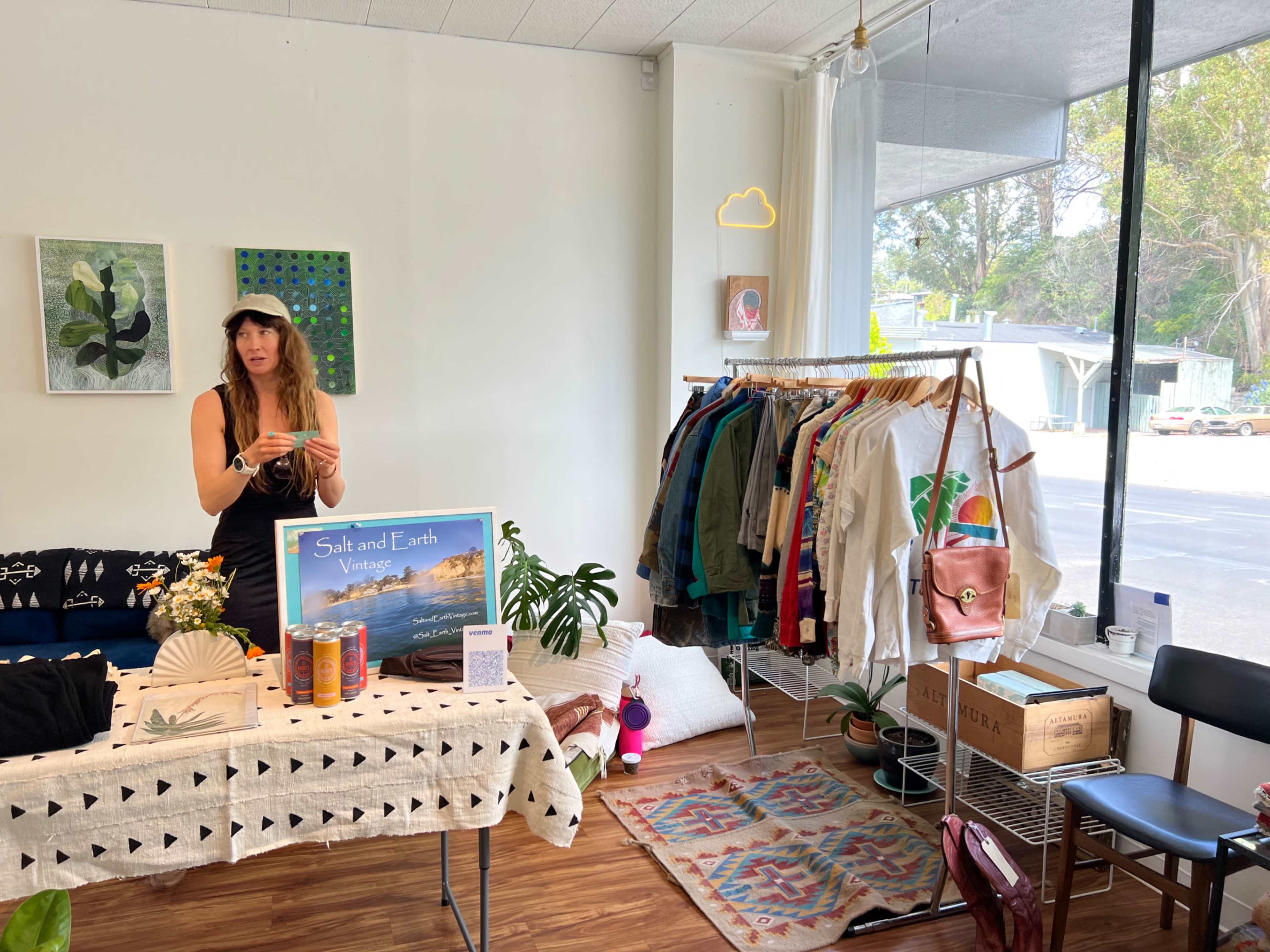 A woman stands behind a display table in a vintage clothing shop, with racks of colorful garments and decorative items surrounding her.