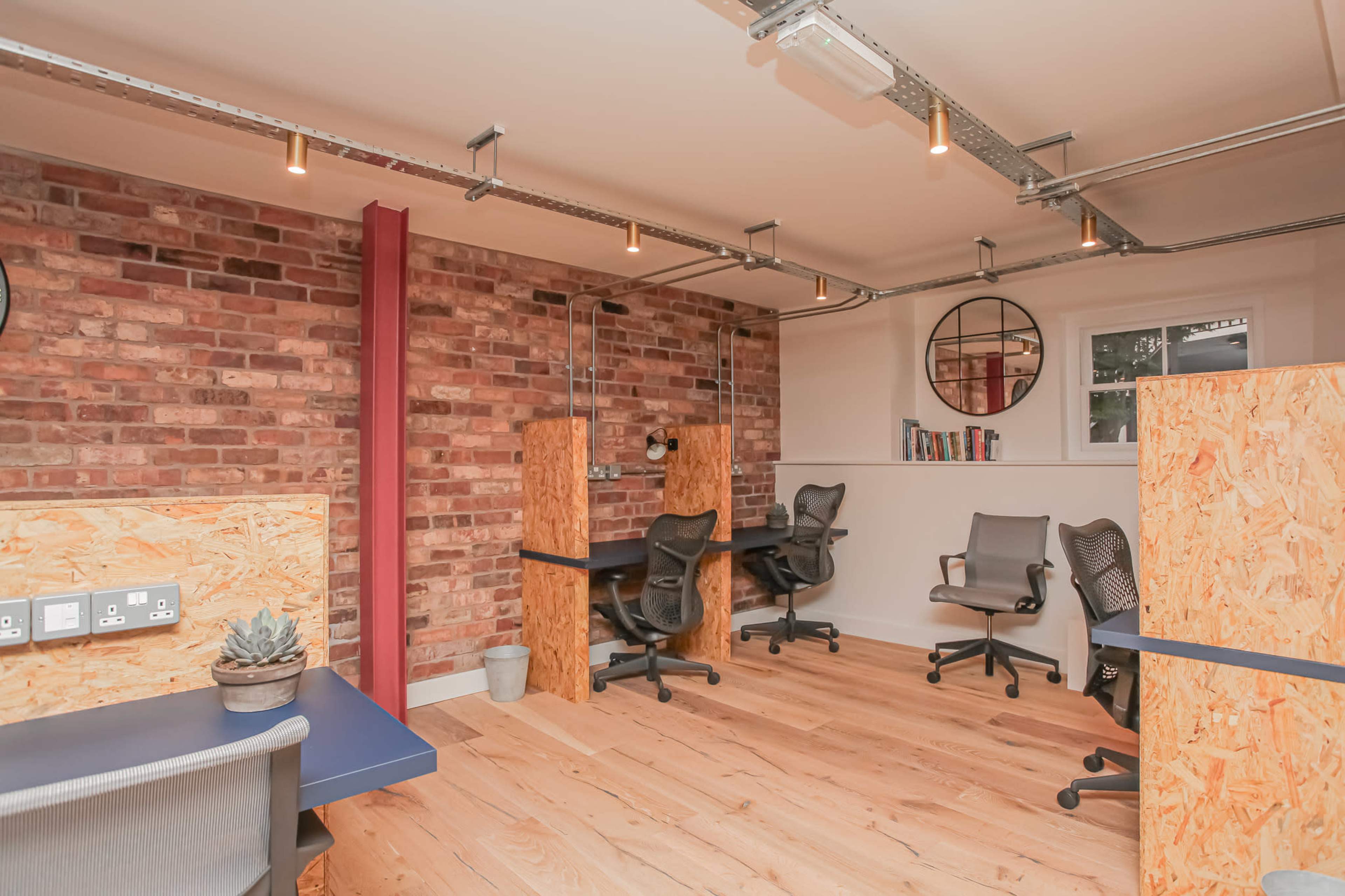The image shows a modern office space with wooden desks and office chairs, featuring industrial-style brick walls and overhead lighting.