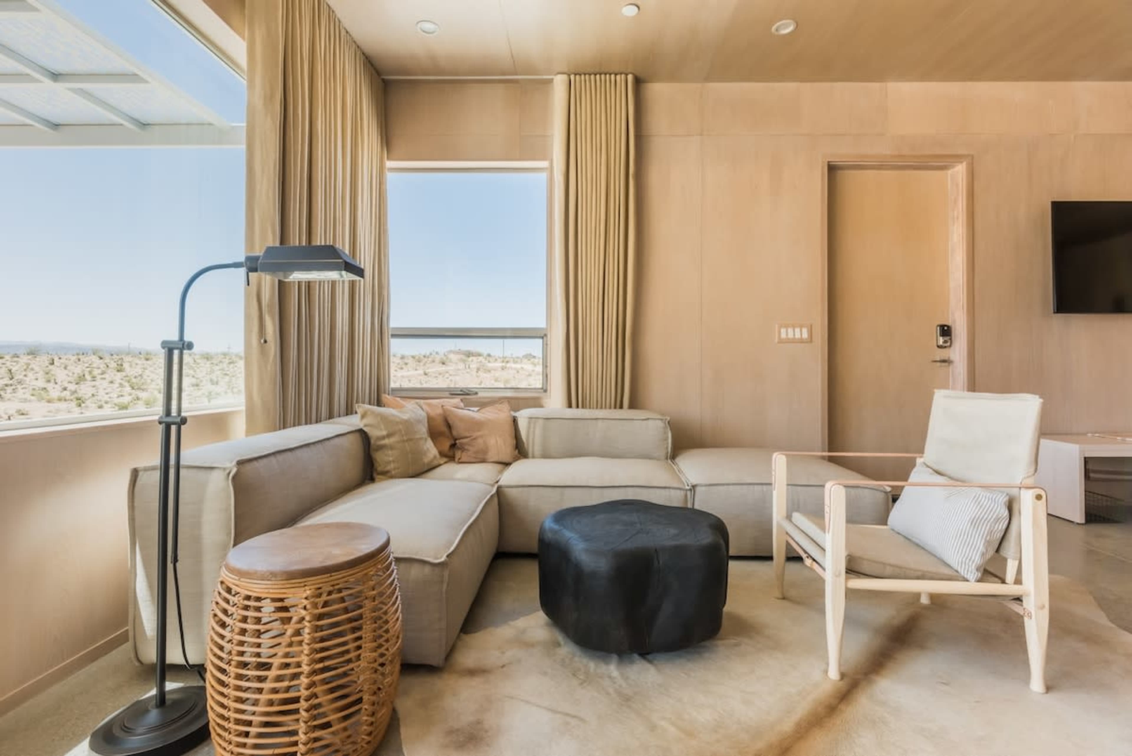 The image depicts a modern living room with a sectional sofa, a circular black table, a wooden accent chair, and large windows showcasing a desert landscape.