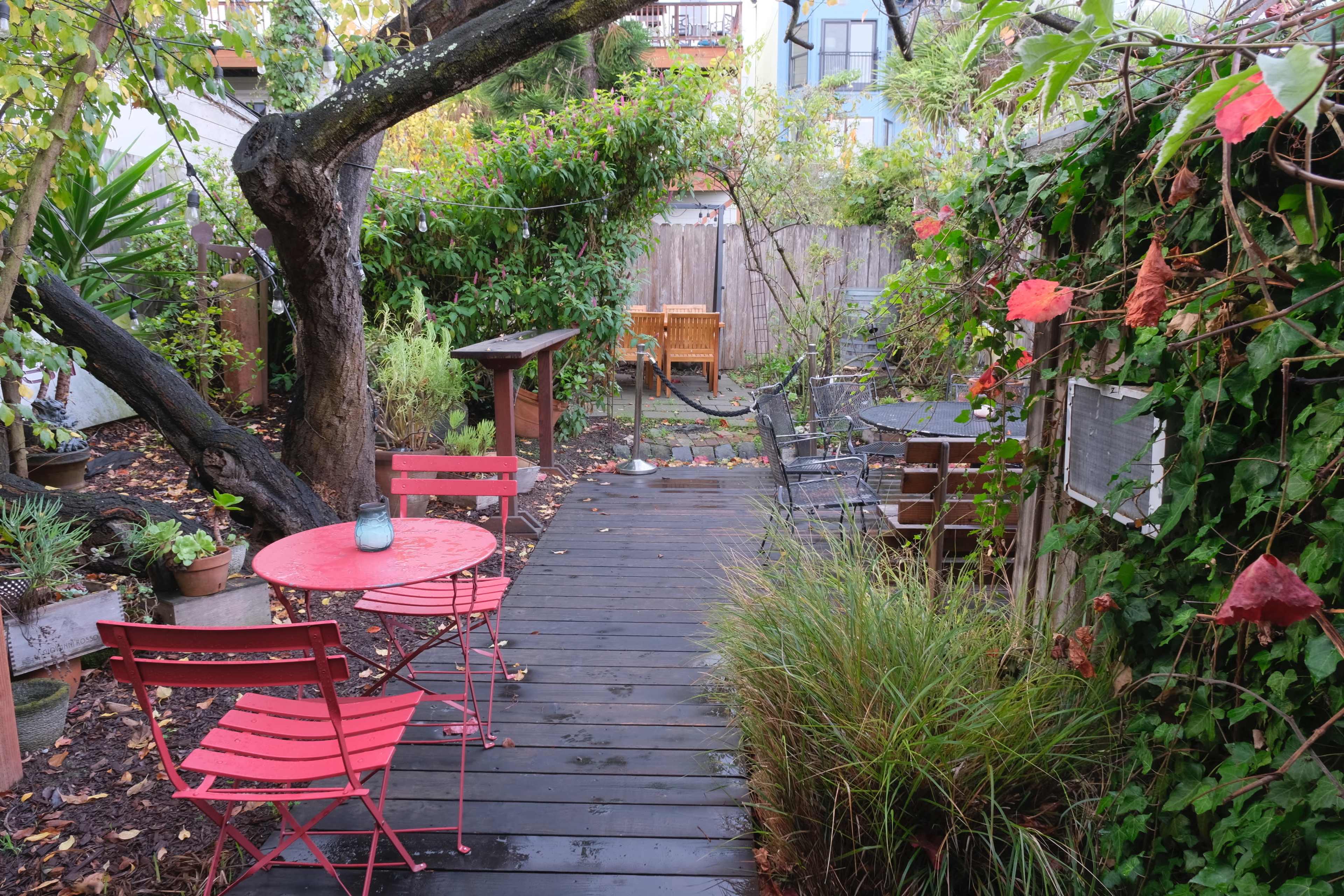 A narrow wooden pathway leads through a garden with potted plants, red chairs and tables, and a wooden seating area in the background.
