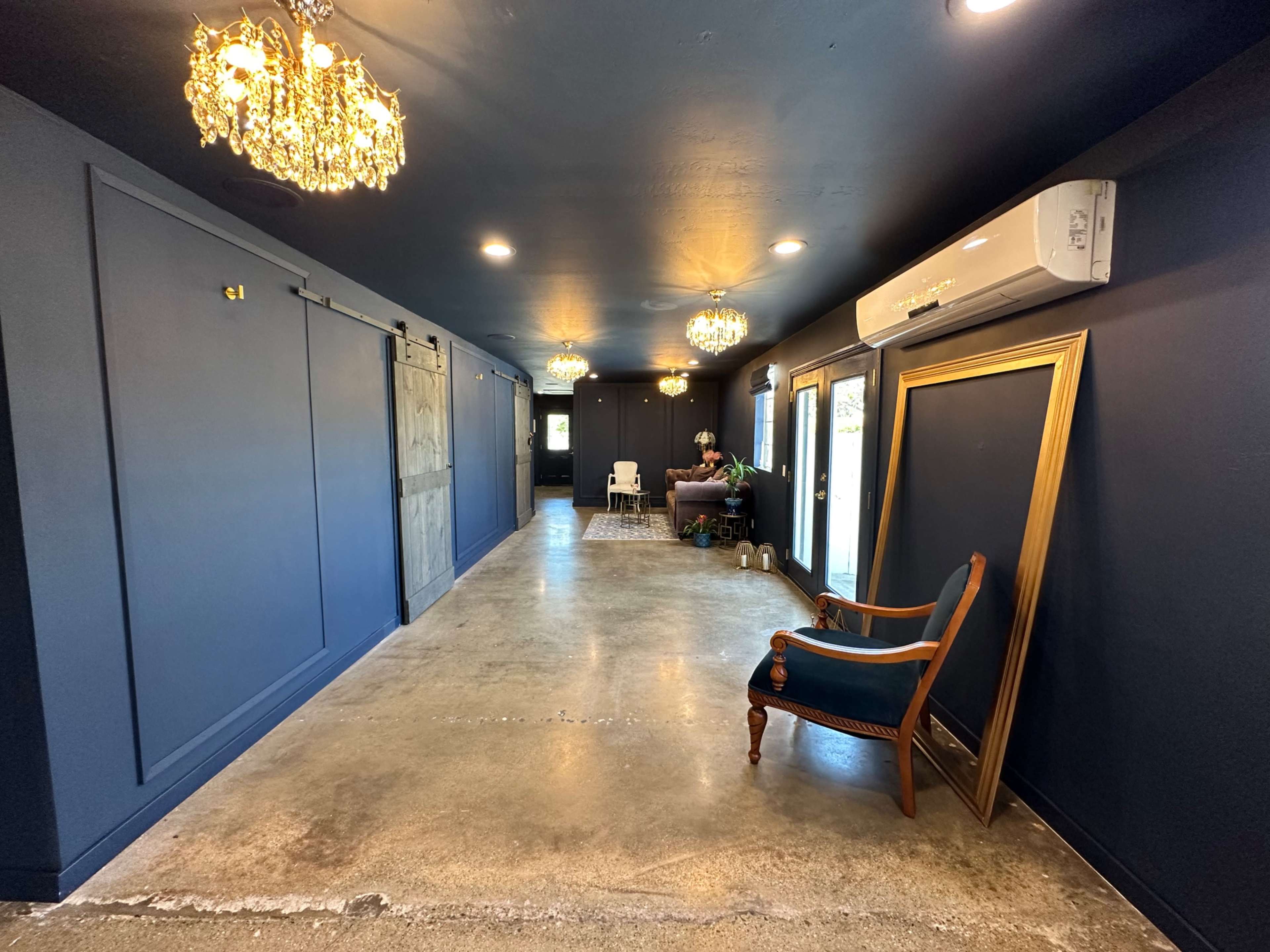 The image shows a narrow hallway with dark walls, illuminated by decorative chandeliers, featuring a large framed mirror and a vintage armchair near one side.