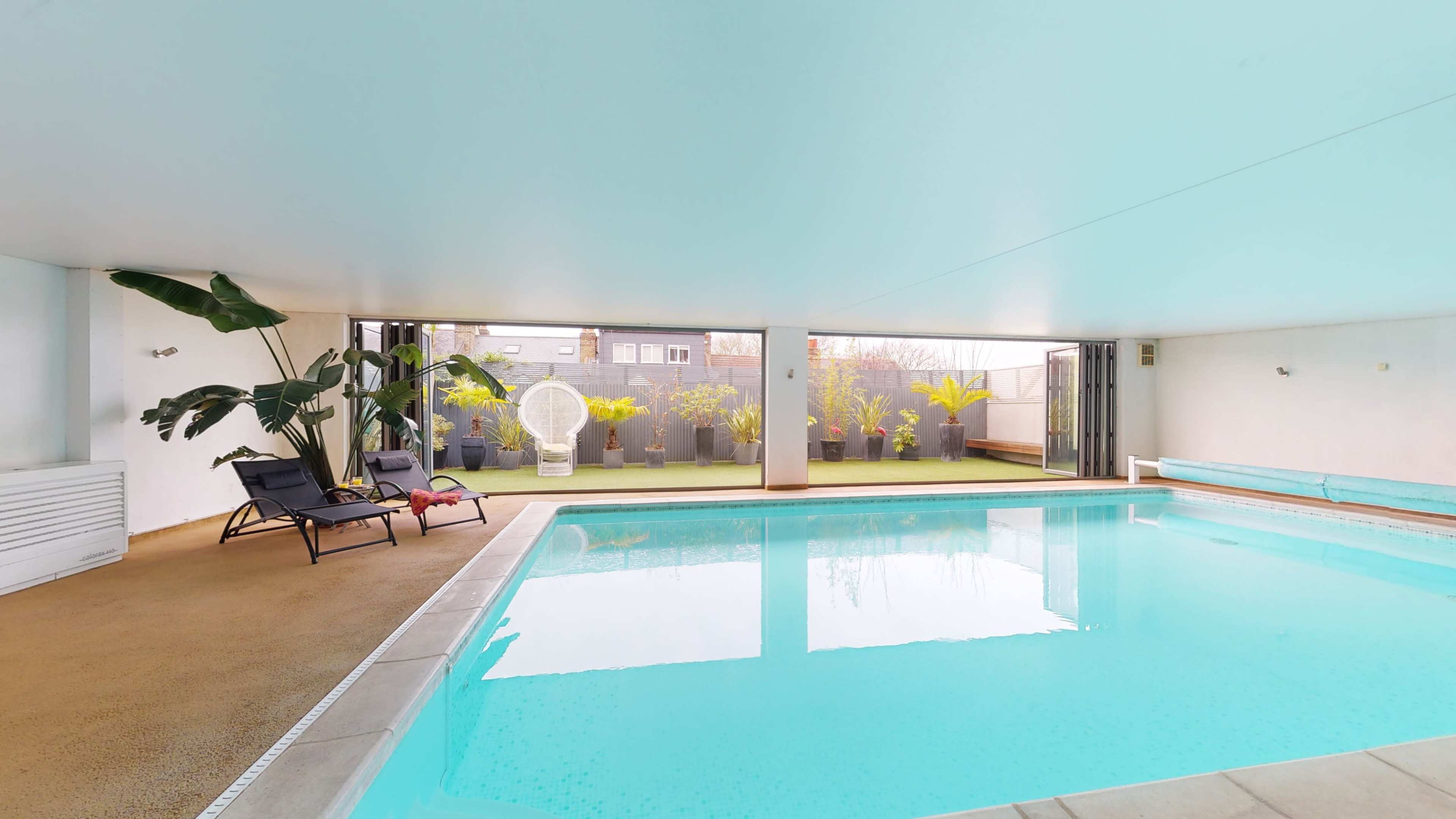 The image shows an indoor swimming pool area with large windows overlooking a small garden, surrounded by lounge chairs and indoor plants.