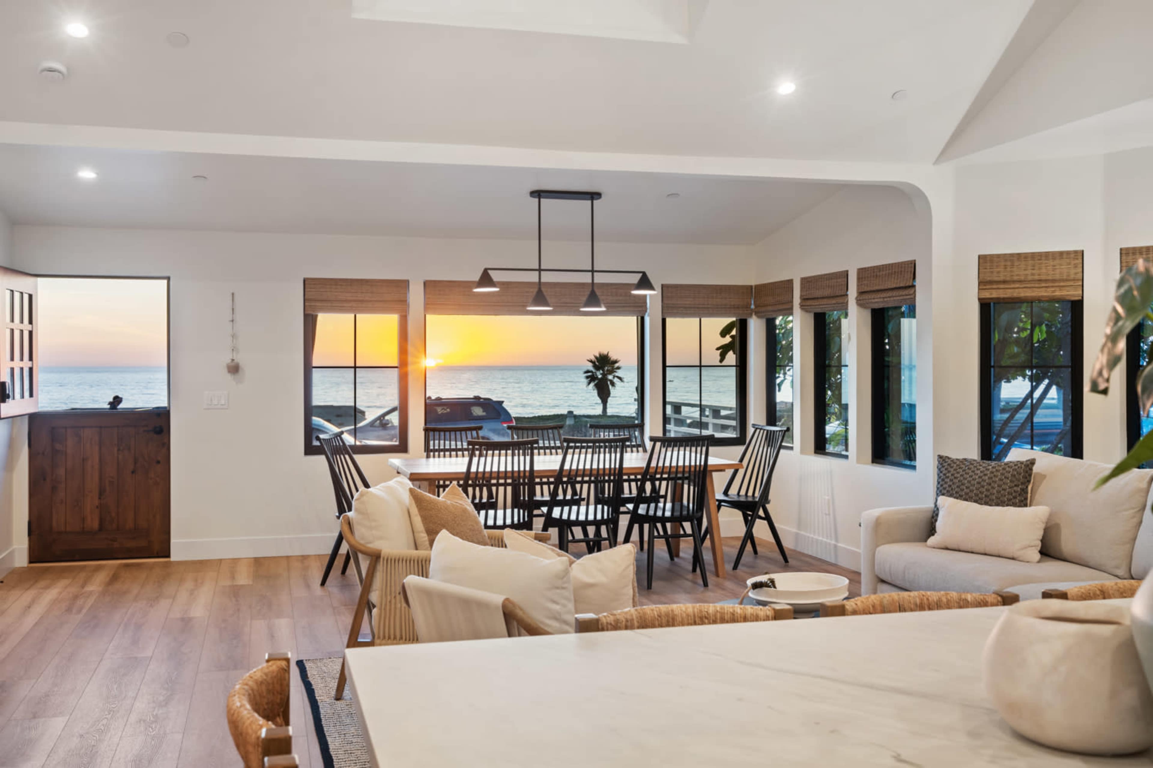 The room features large windows that offer a view of the sunset over the ocean, with a dining table and seating arranged for socializing.