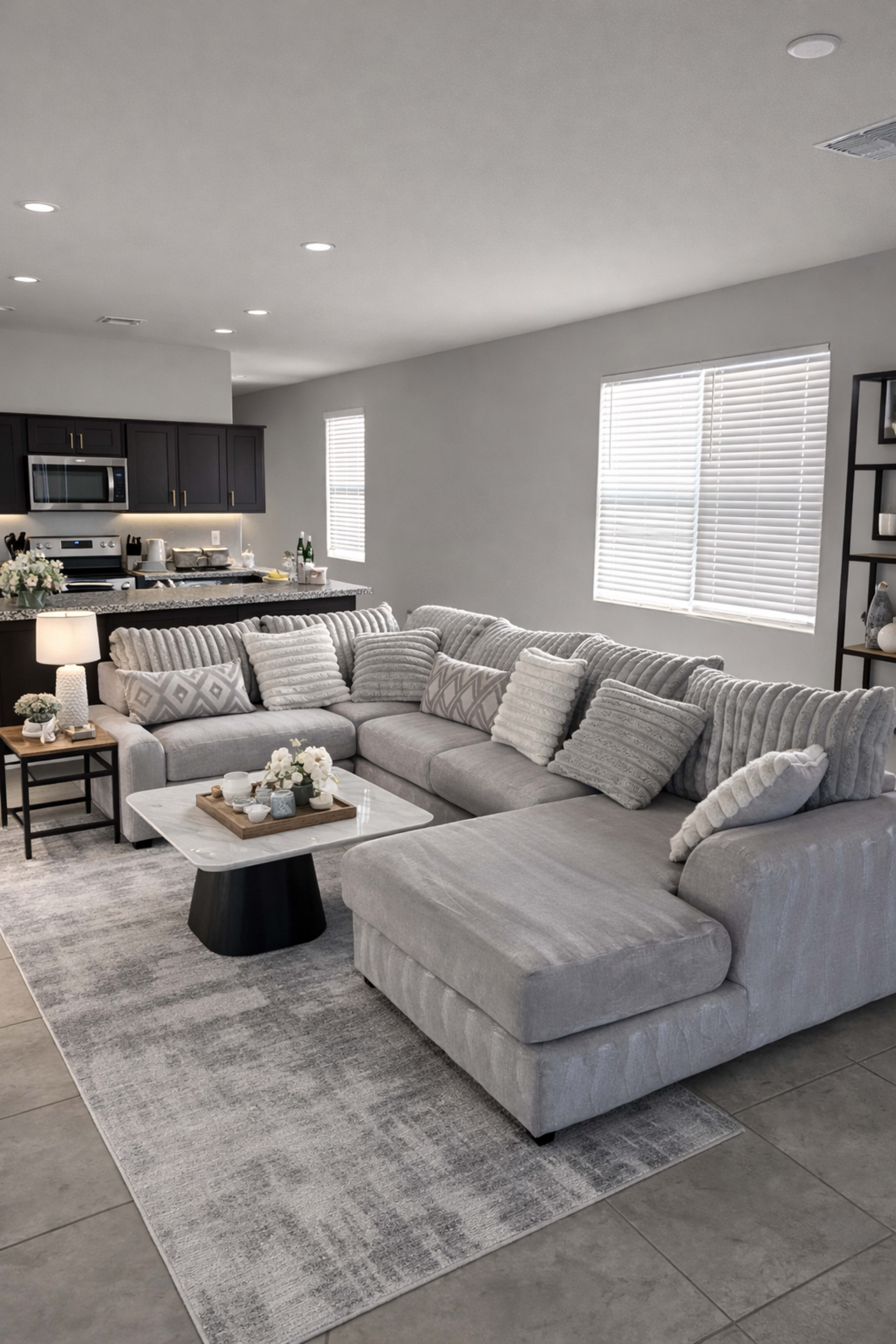 The living room features a large gray sectional sofa, a white coffee table, and an open view of a modern kitchen area with dark cabinets.