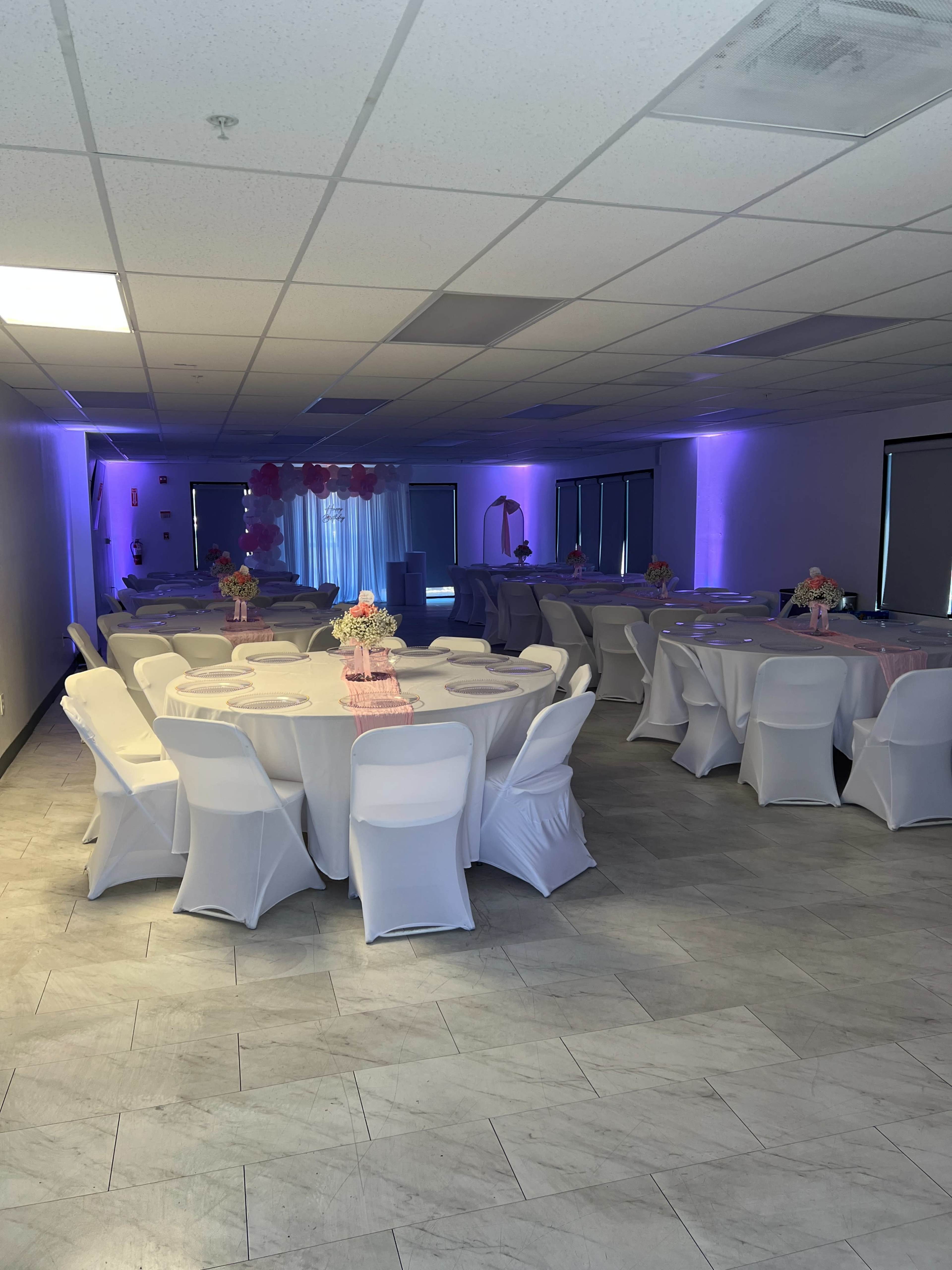 The image shows a decorated event space with white tables and chairs set up, featuring pink floral centerpieces and purple lighting.