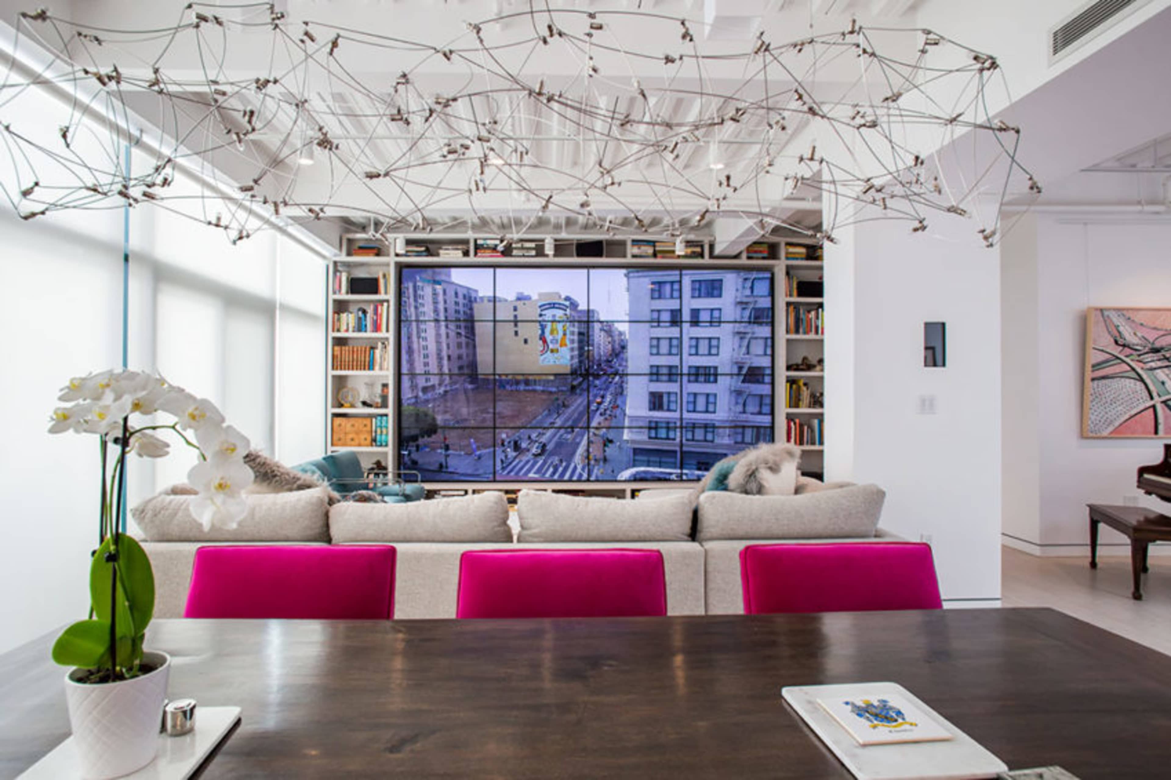 A modern living room features a large television mounted on a white wall, surrounded by bookshelves, with a couch adorned with pink cushions and a dining table in the foreground.