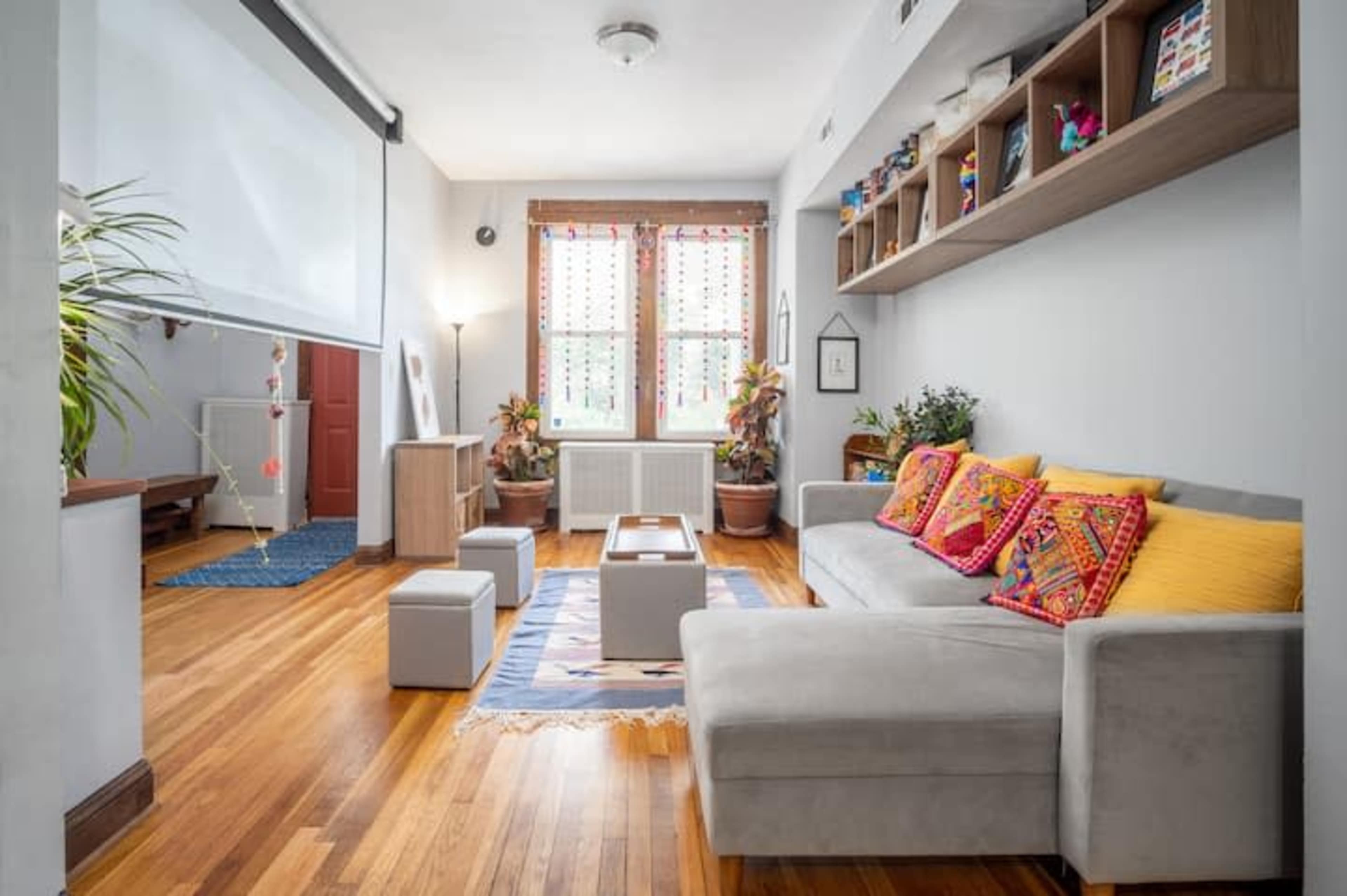 A living room features a gray sofa with colorful cushions, a wooden coffee table, and large windows adorned with decorative curtains.