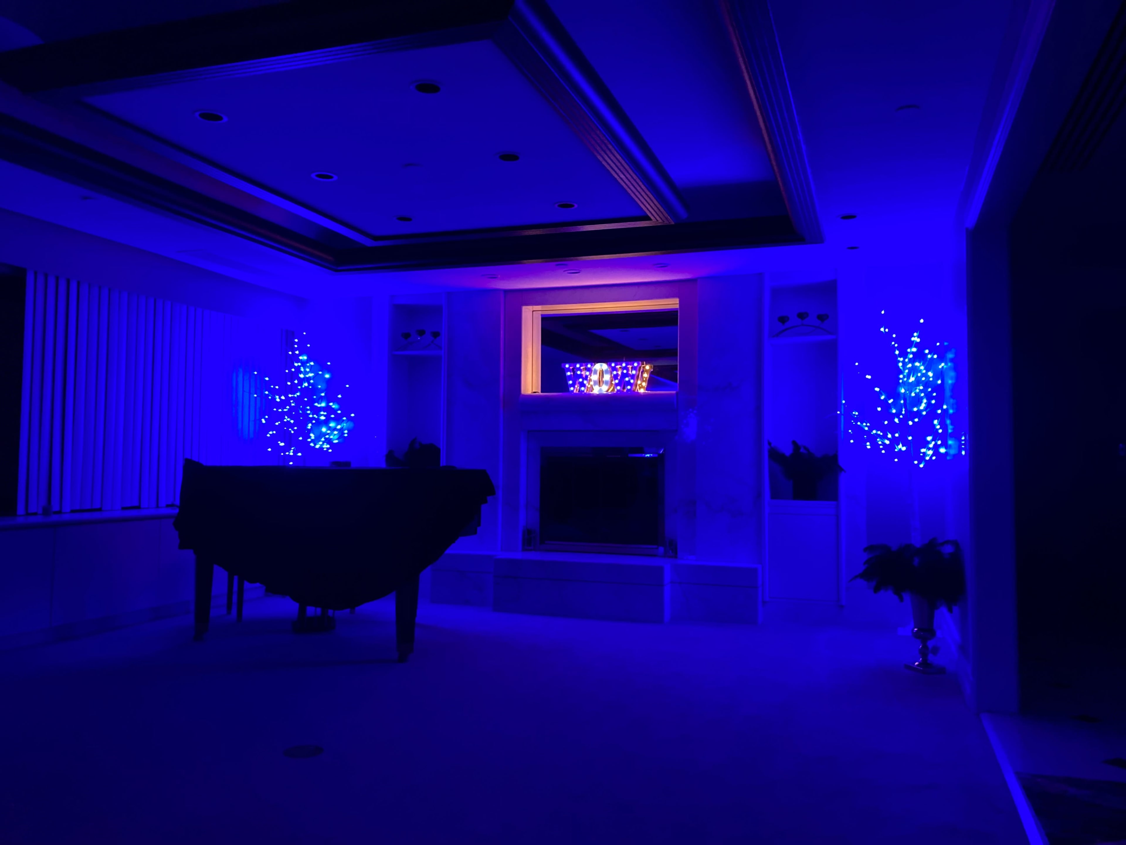 A dimly lit room features a pool table covered with a black cloth, blue LED-lit trees, and a fireplace surrounded by glowing decorations.