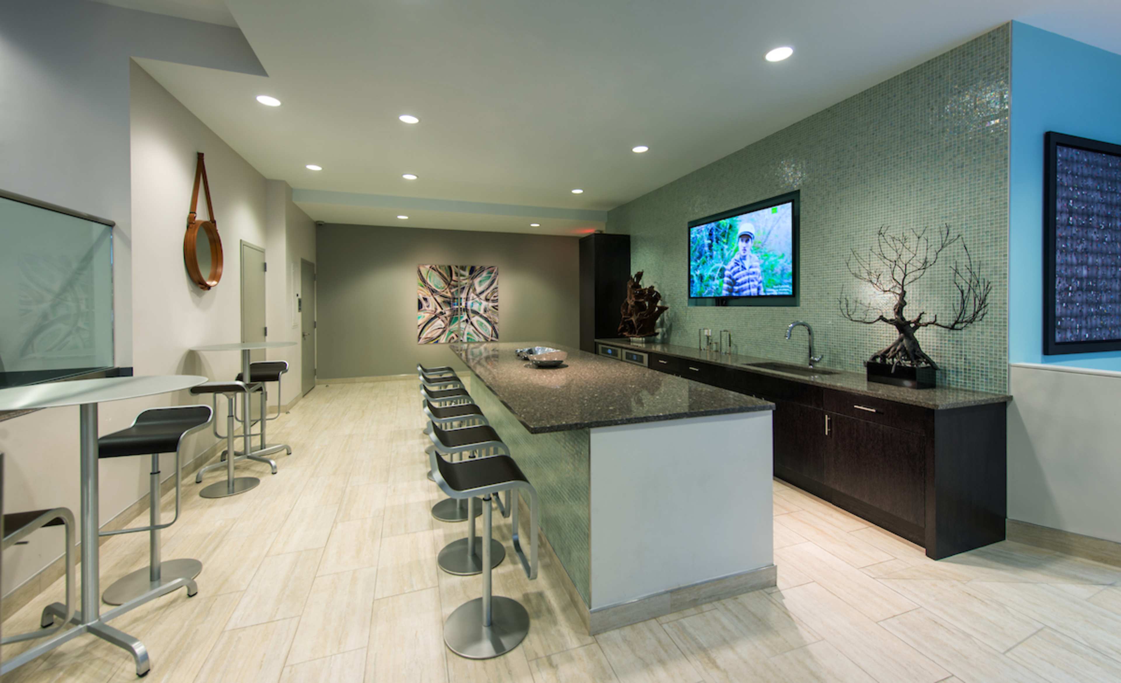 A modern kitchenette with a long granite counter, bar stools, a television on the wall, and artwork displayed on the adjacent wall.