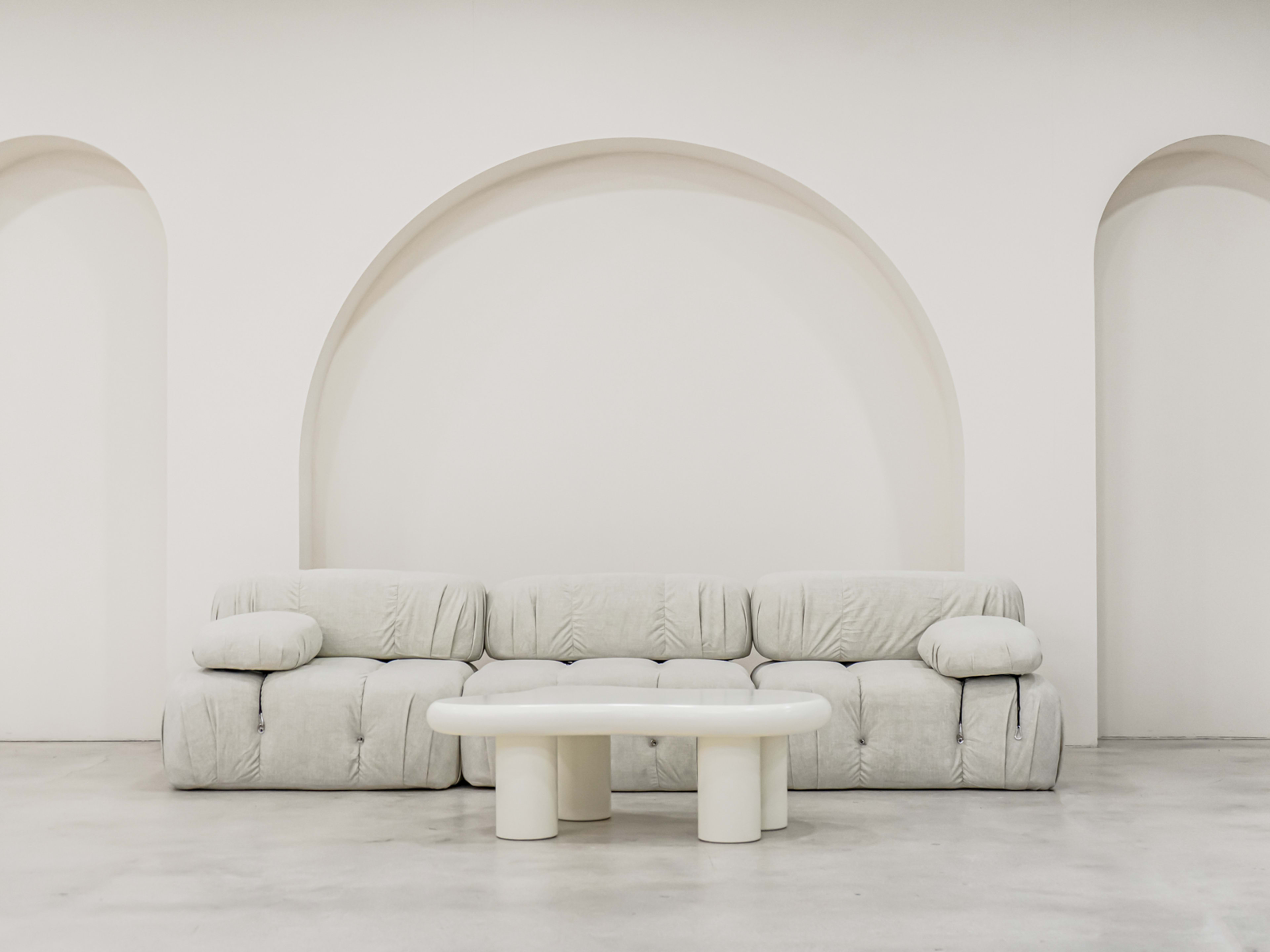 The image shows a light-colored, upholstered sofa situated in front of a white wall with an arched recess, accompanied by a round, white coffee table.