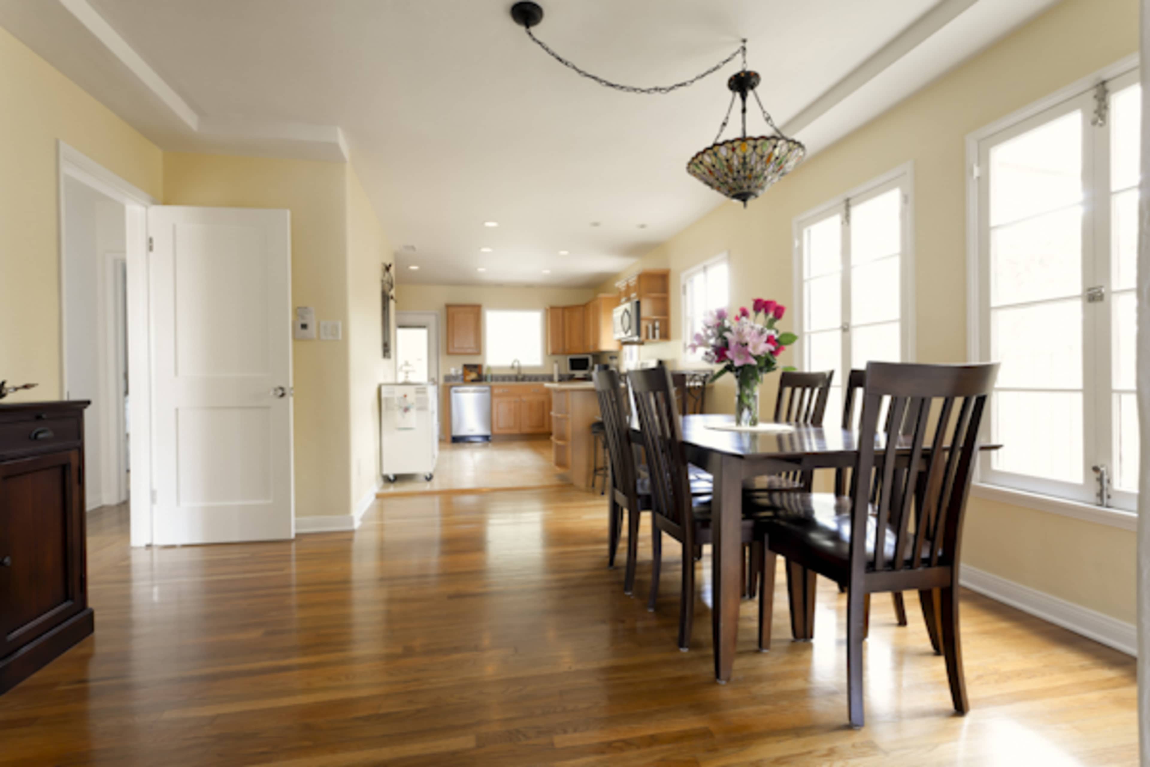 A dining area features a wooden table with six chairs and a chandelier, leading into a kitchen with wooden cabinets and multiple windows.