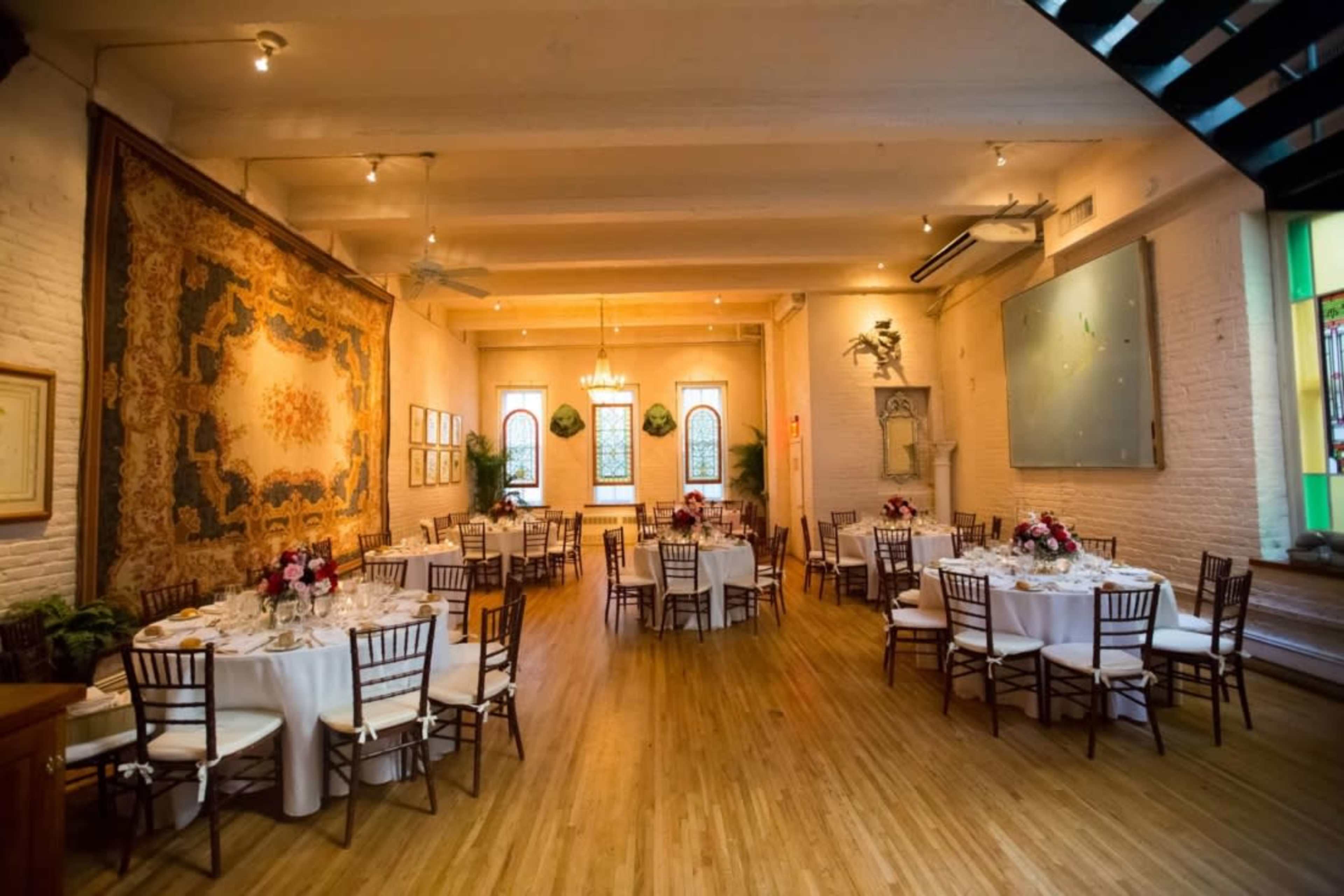 The image shows a spacious event room with wooden floors, decorated tables set for a meal, and large windows allowing natural light to filter in.
