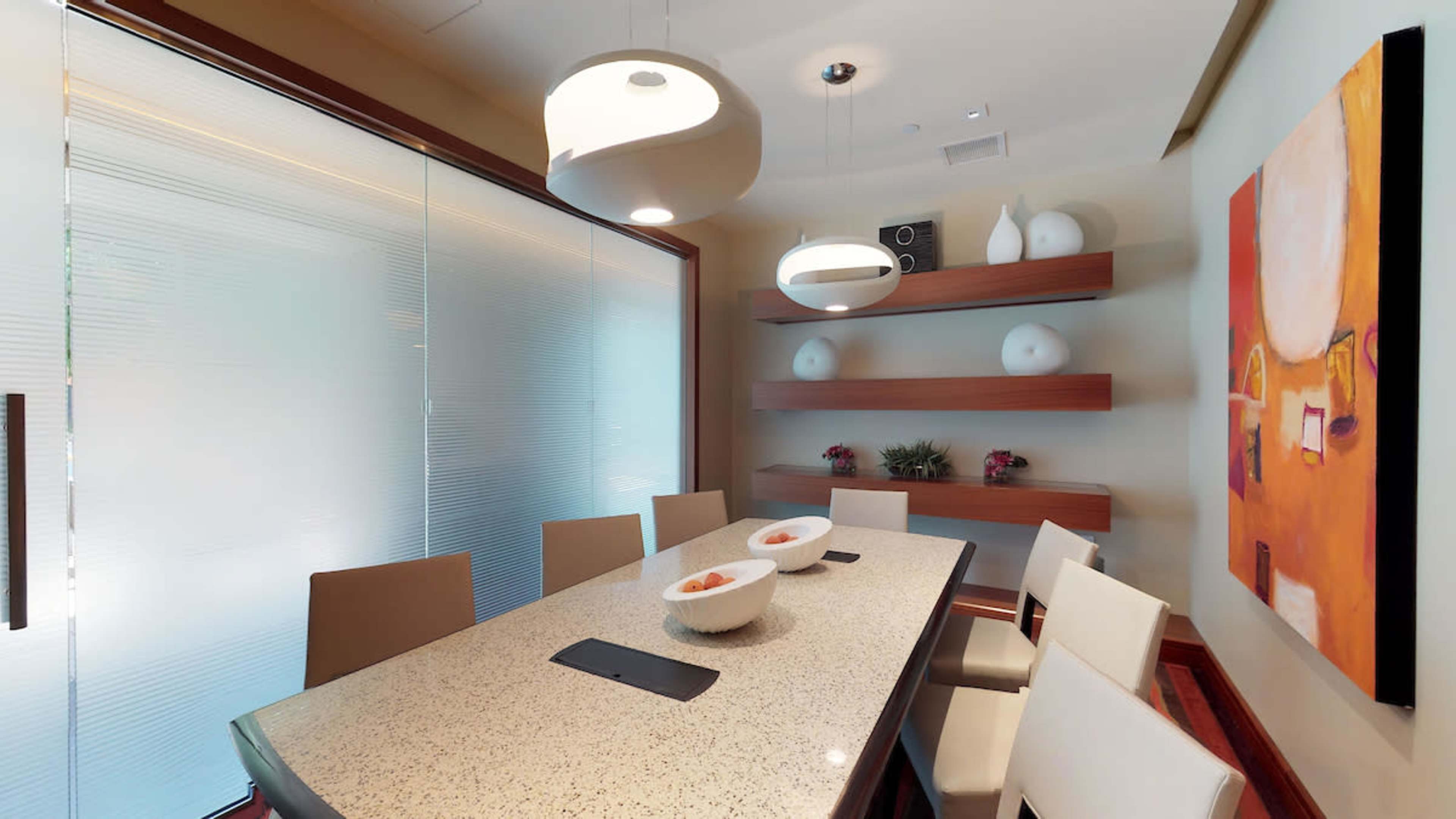 A modern dining area features a long table with eight chairs, illuminated by hanging light fixtures, and is adorned with minimalist decor and a colorful wall painting.