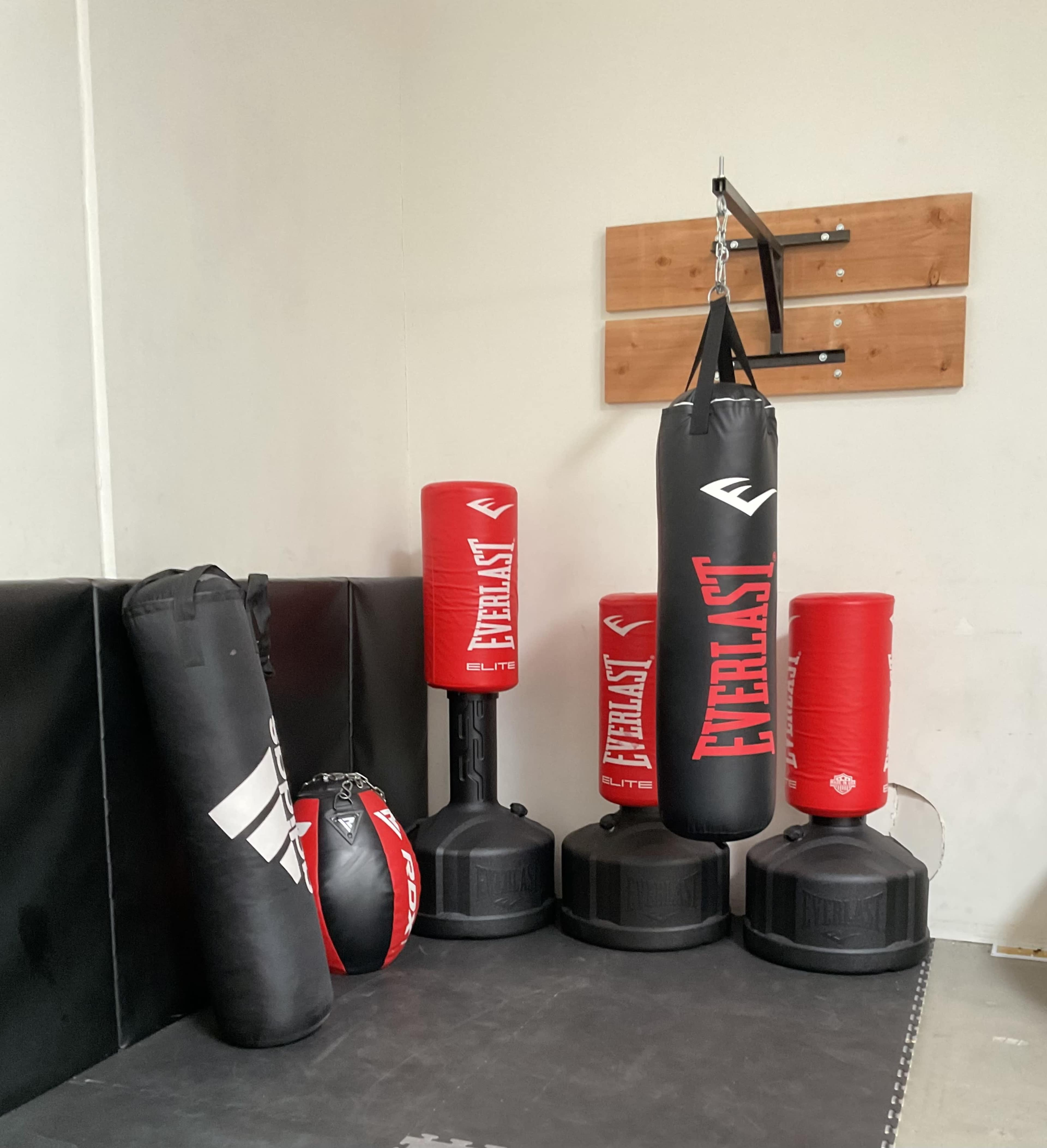The image shows a corner of a gym with various boxing equipment, including standing bags, a hanging heavy bag, and training balls arranged neatly against a wall.