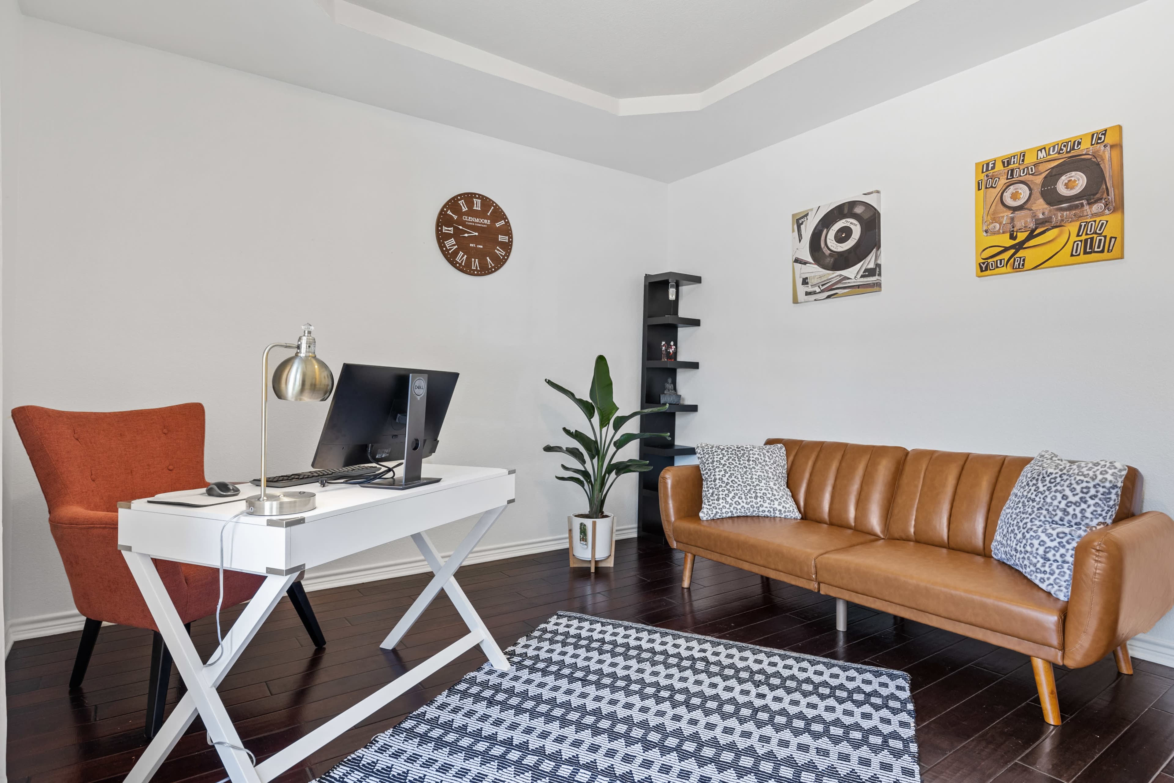 A modern home office with a desk, a computer, an orange chair, a brown leather sofa, and decor including wall art and a clock.