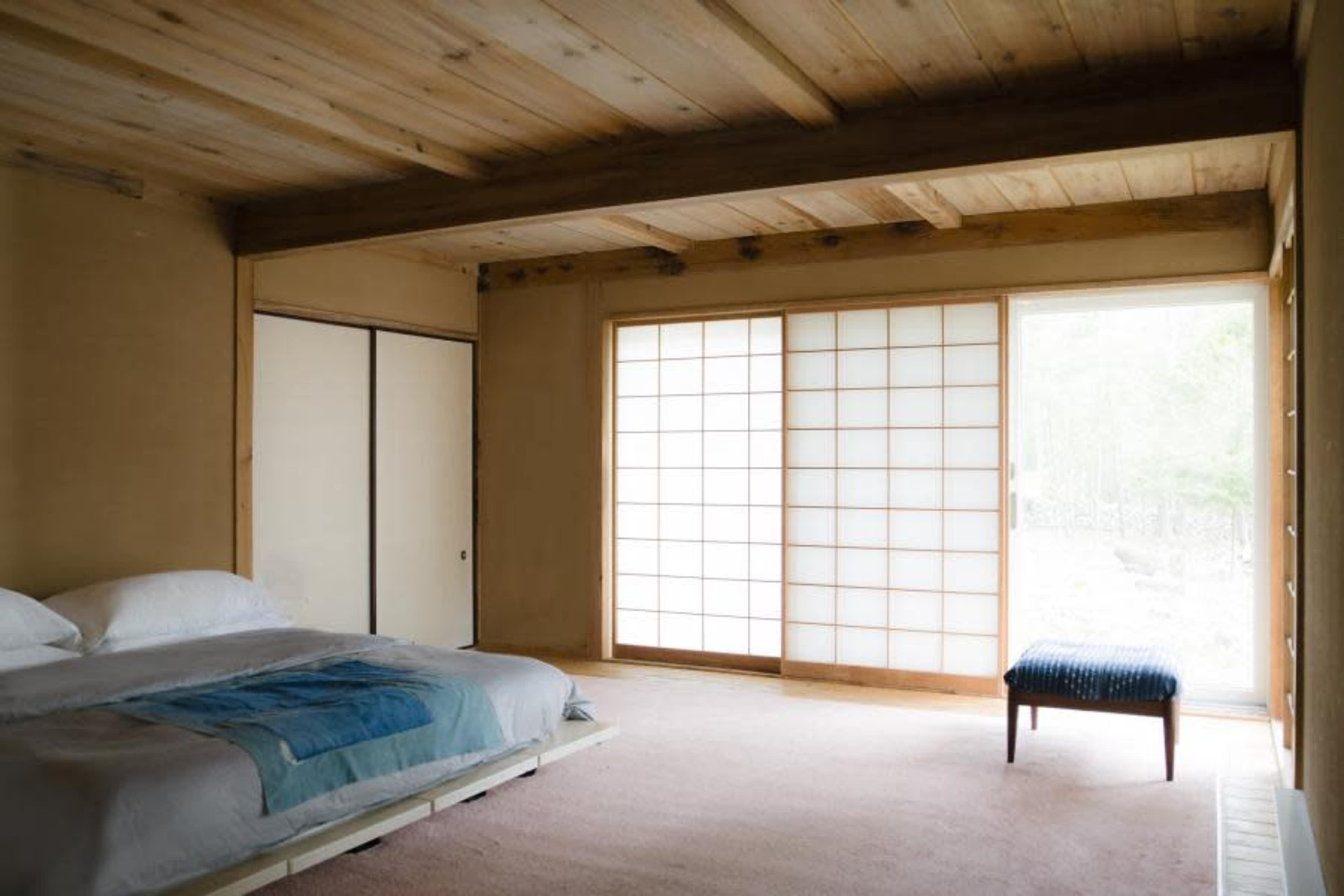 The image shows a minimalist bedroom with a bed and a shoji screen, adjacent to a sliding glass door that opens to a natural view.