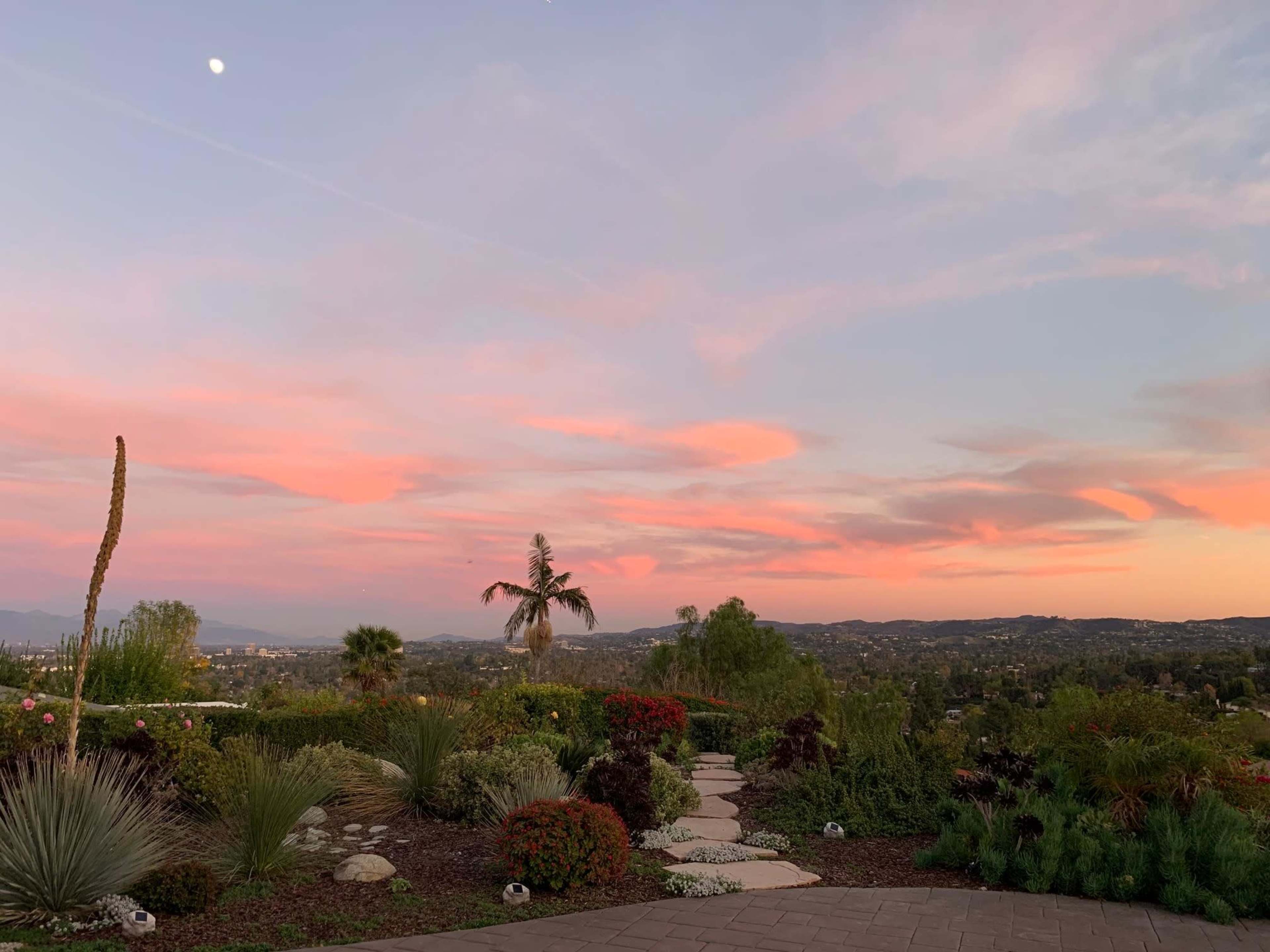 A pathway winds through a landscaped garden under a colorful sunset sky with distant mountains.