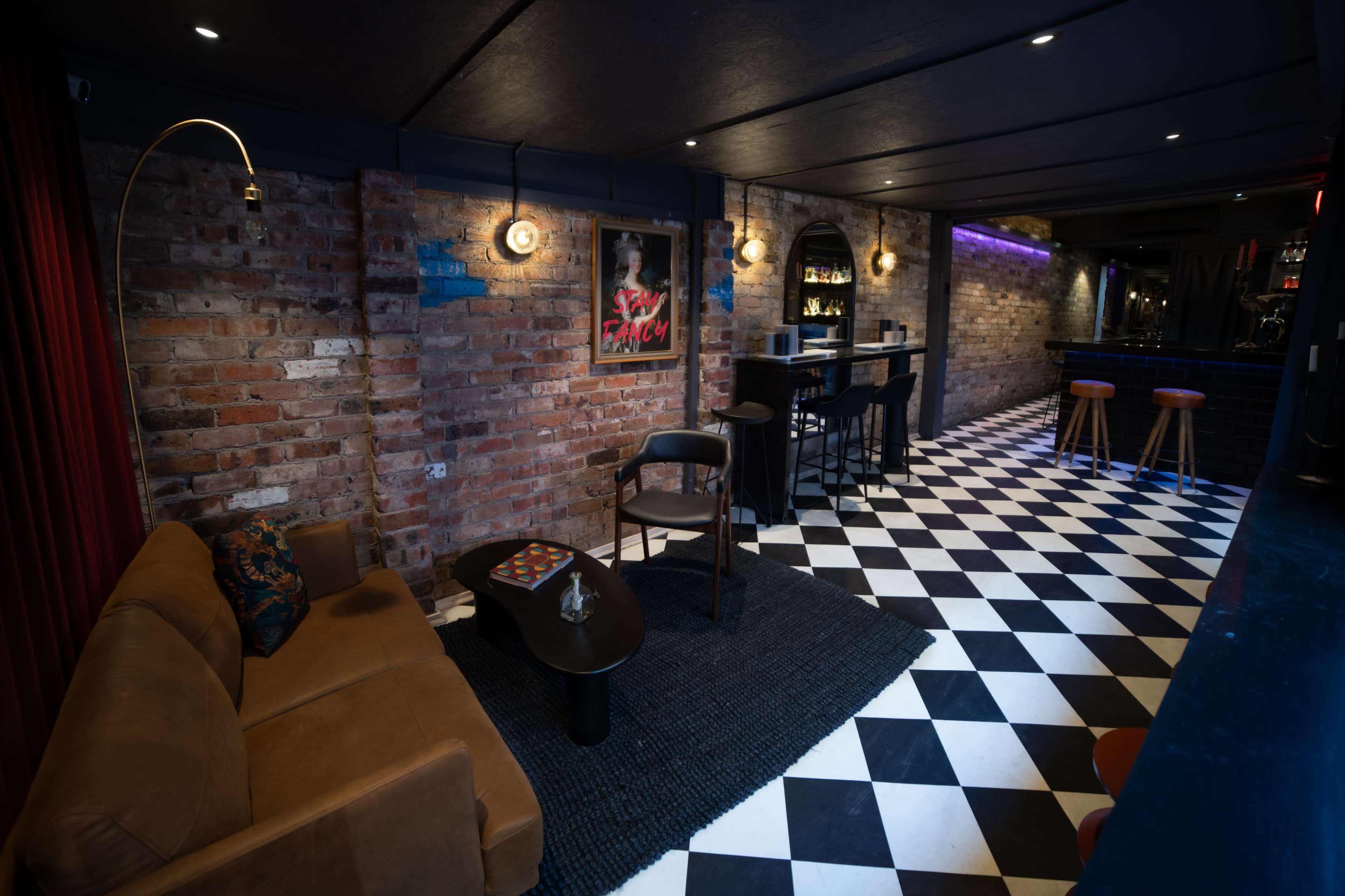The image shows a stylish interior of a bar or lounge featuring exposed brick walls, a checkered floor, and various seating arrangements.