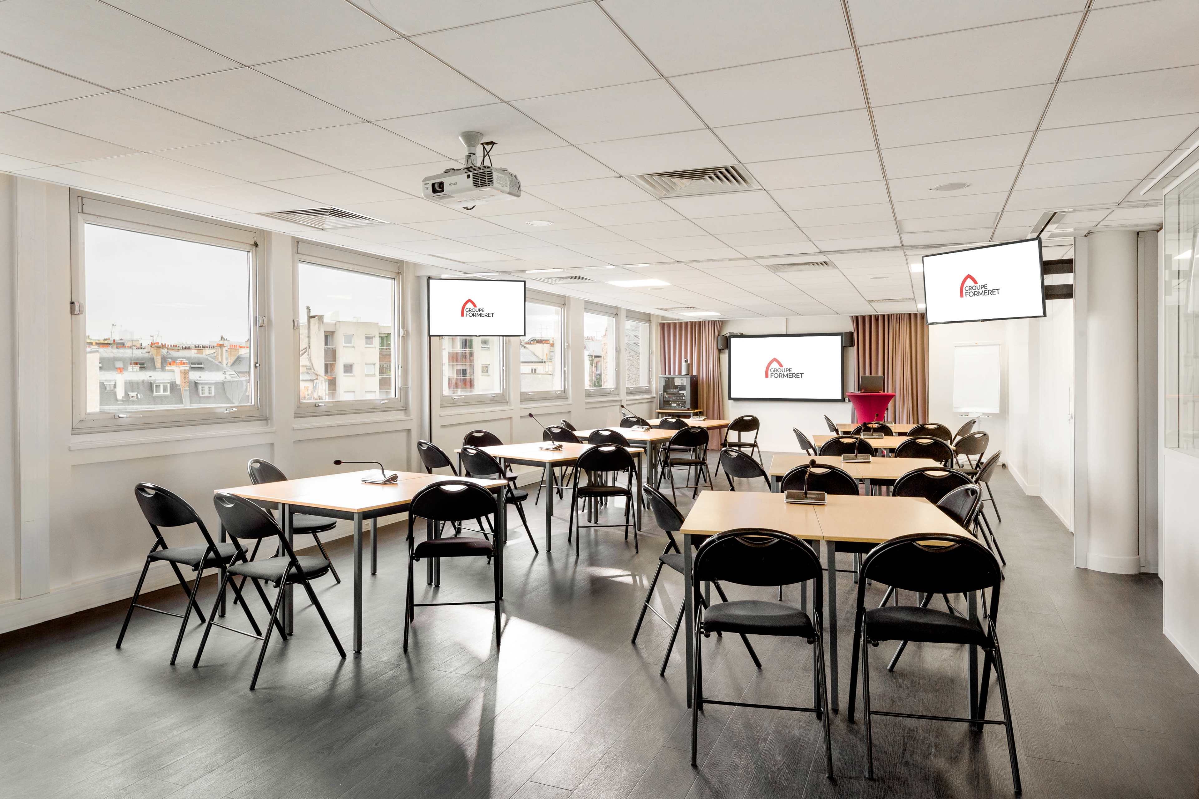A modern classroom setup features several tables and chairs arranged for a presentation, with large screens displaying a logo on the walls.