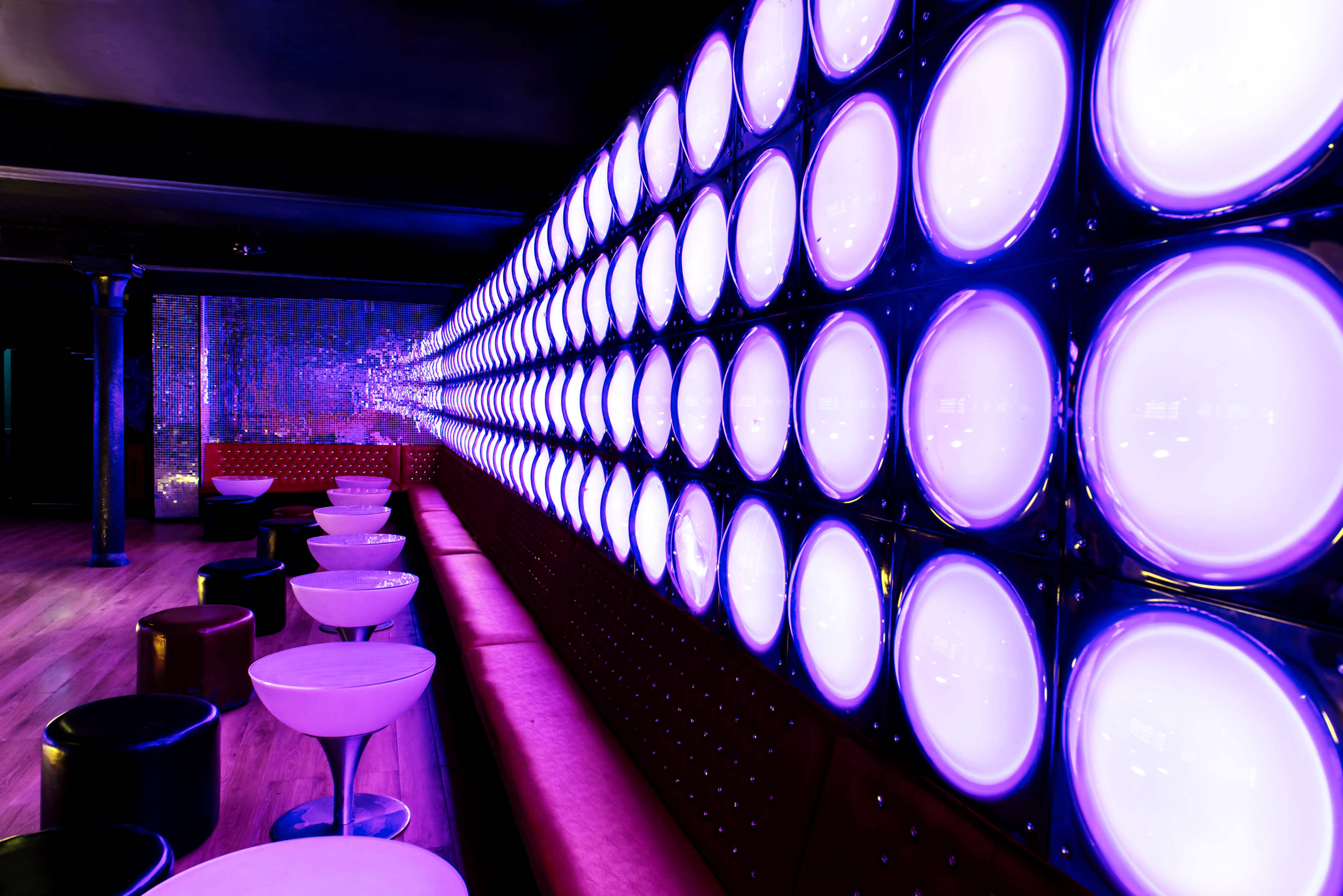 A stylish bar interior with a row of circular purple-lit decorative panels and white stools along a red upholstered seating area.