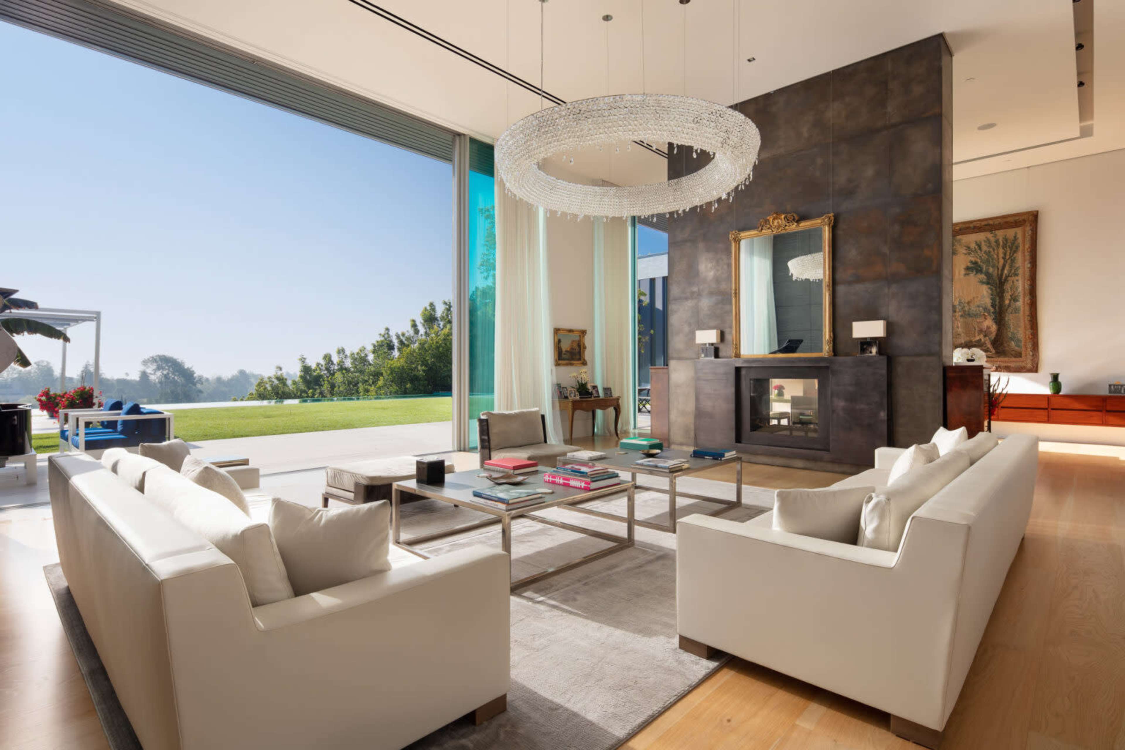 A modern living room with large windows, a contemporary chandelier, and white sofas arranged around a coffee table.