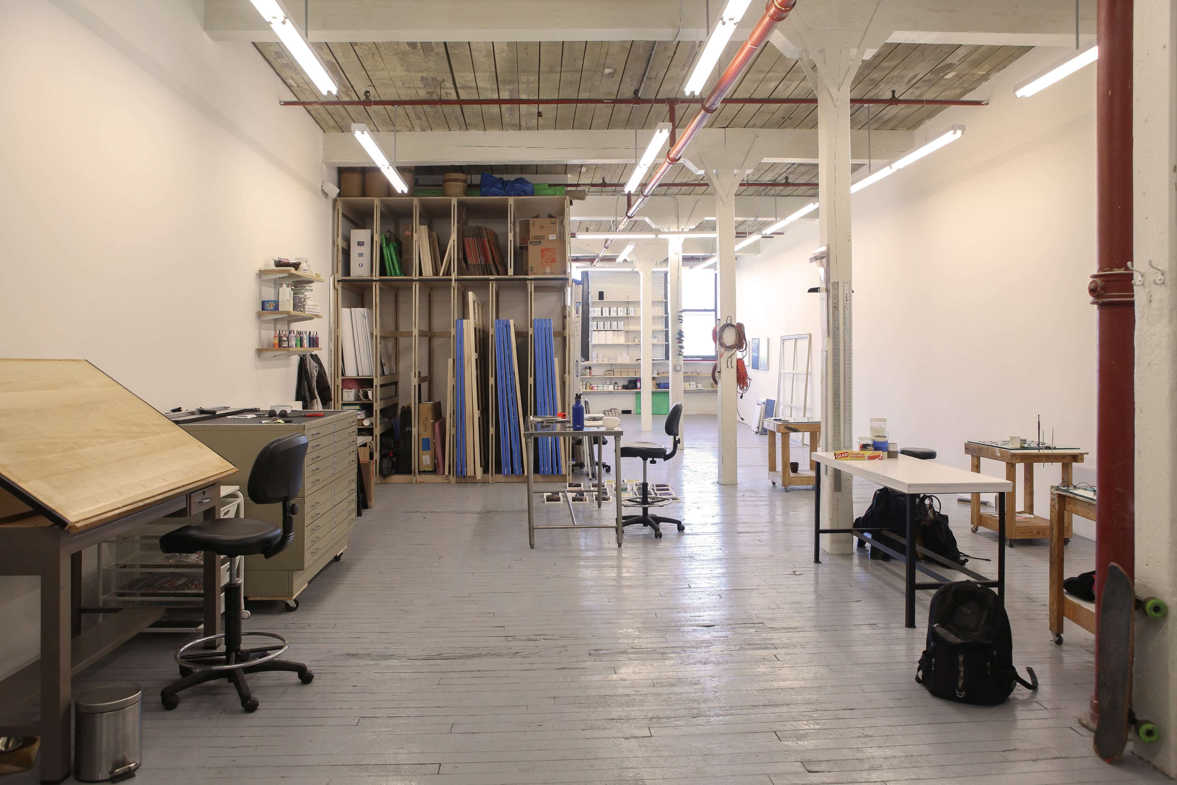 The image shows an open studio space with wooden floors, worktables, art supplies, and shelving filled with materials.