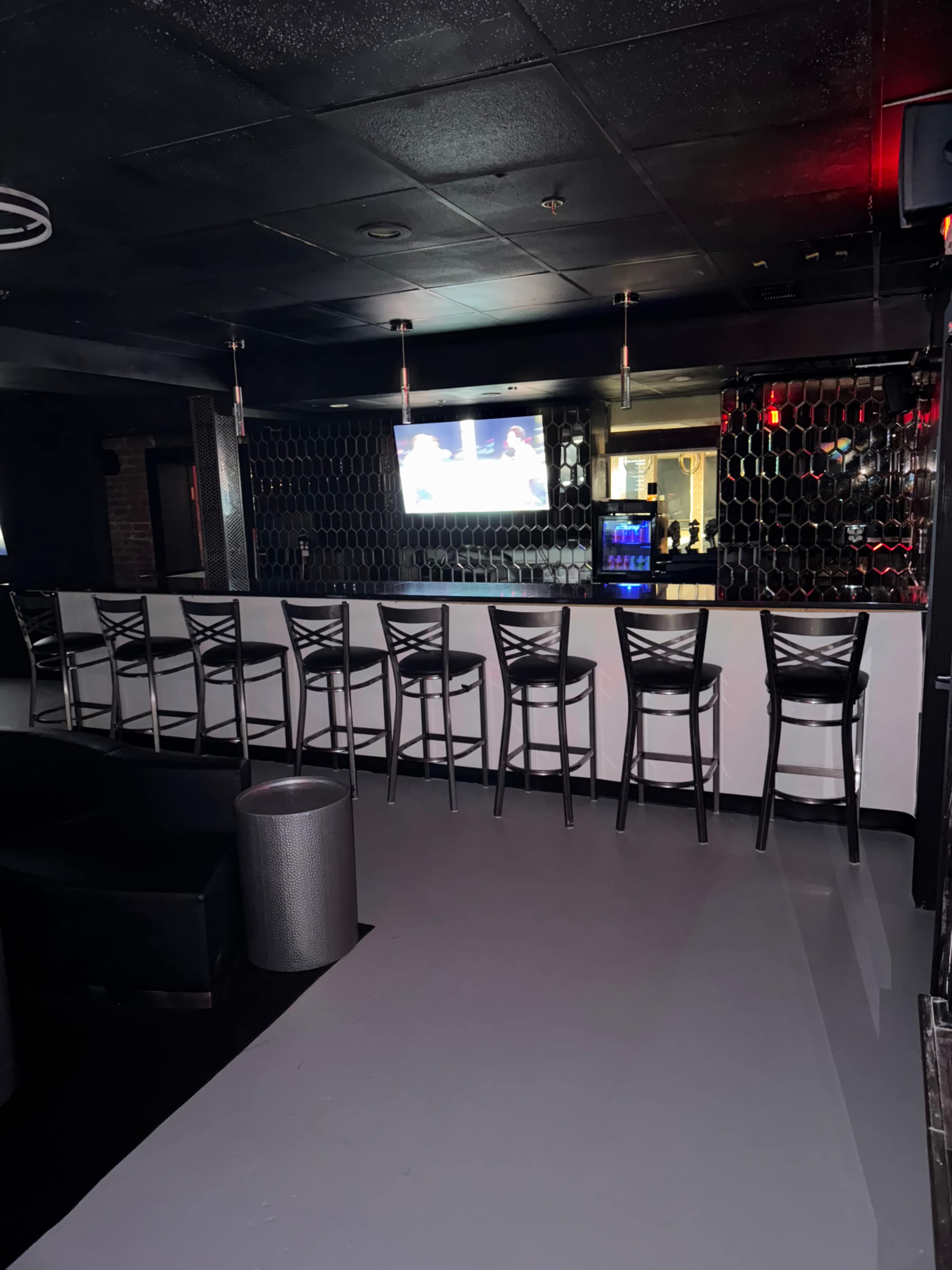 The image shows a dimly lit bar area with a row of black chairs facing a long white counter, and a television mounted on the wall displaying a sports broadcast.