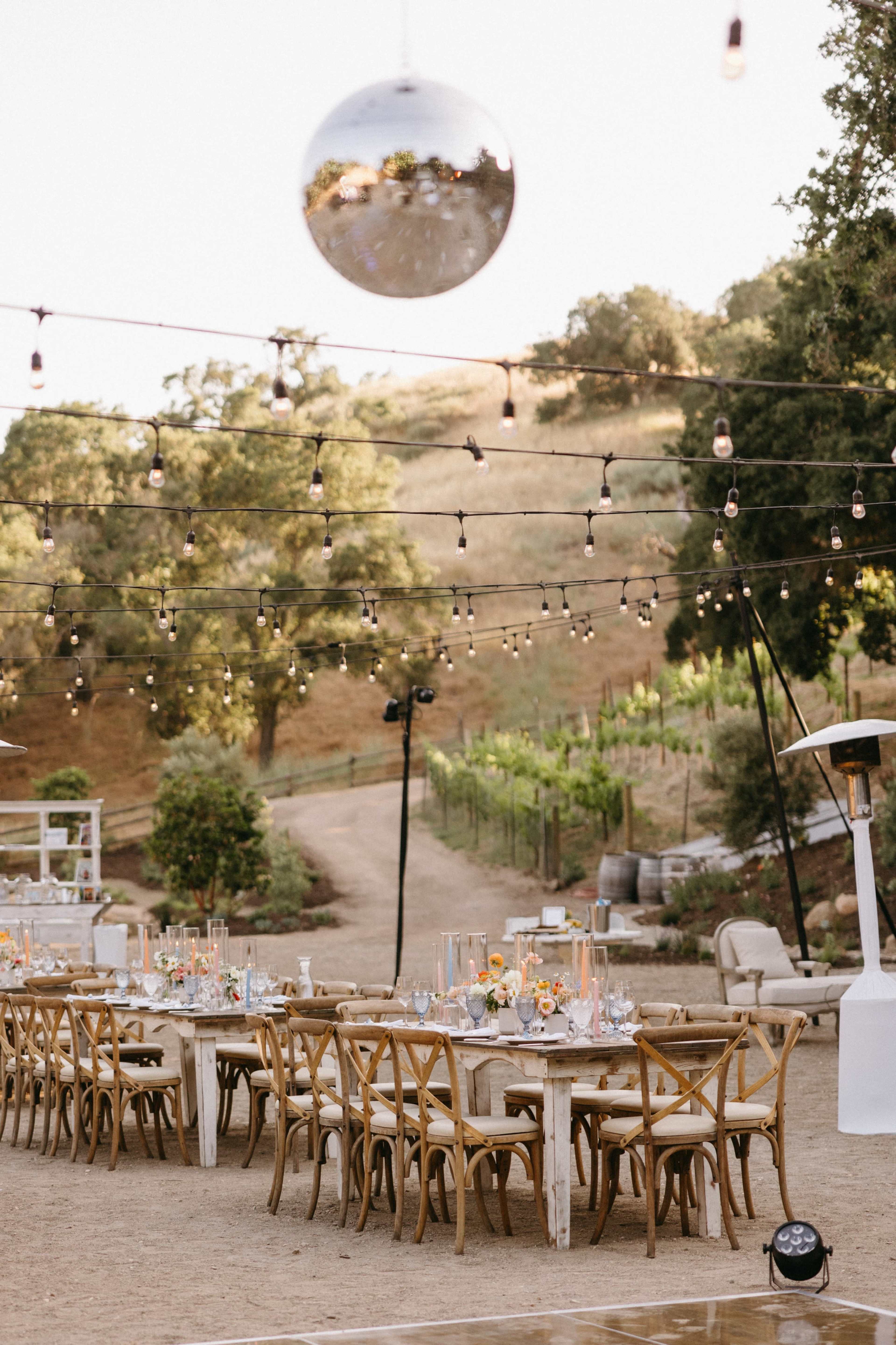 A long, wooden dining table is set outdoors with string lights and a large disco ball overhead, surrounded by vineyards and trees in the background.