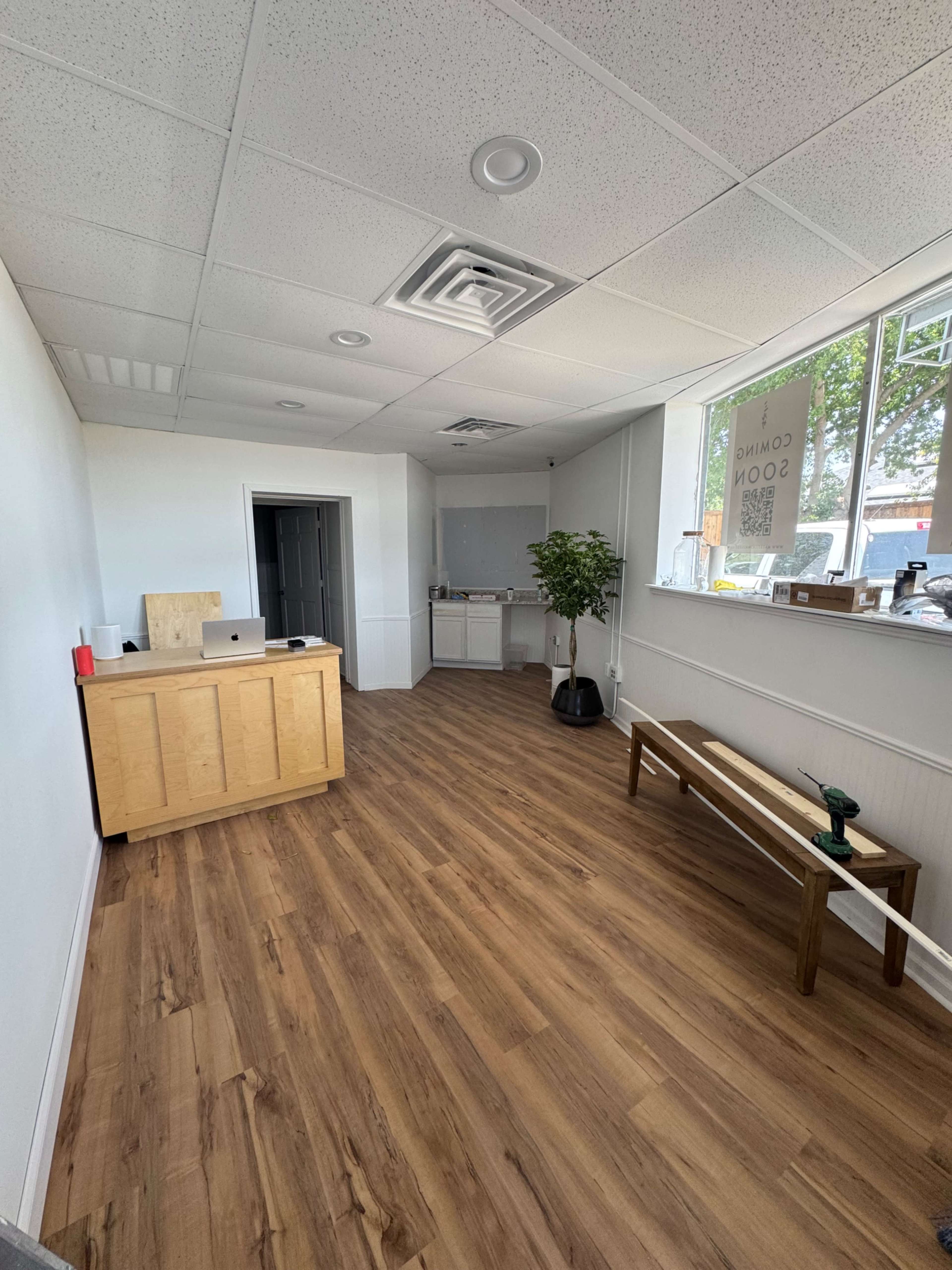 The image shows a vacant office space with a wooden reception desk, a bench, and large windows allowing natural light to enter.
