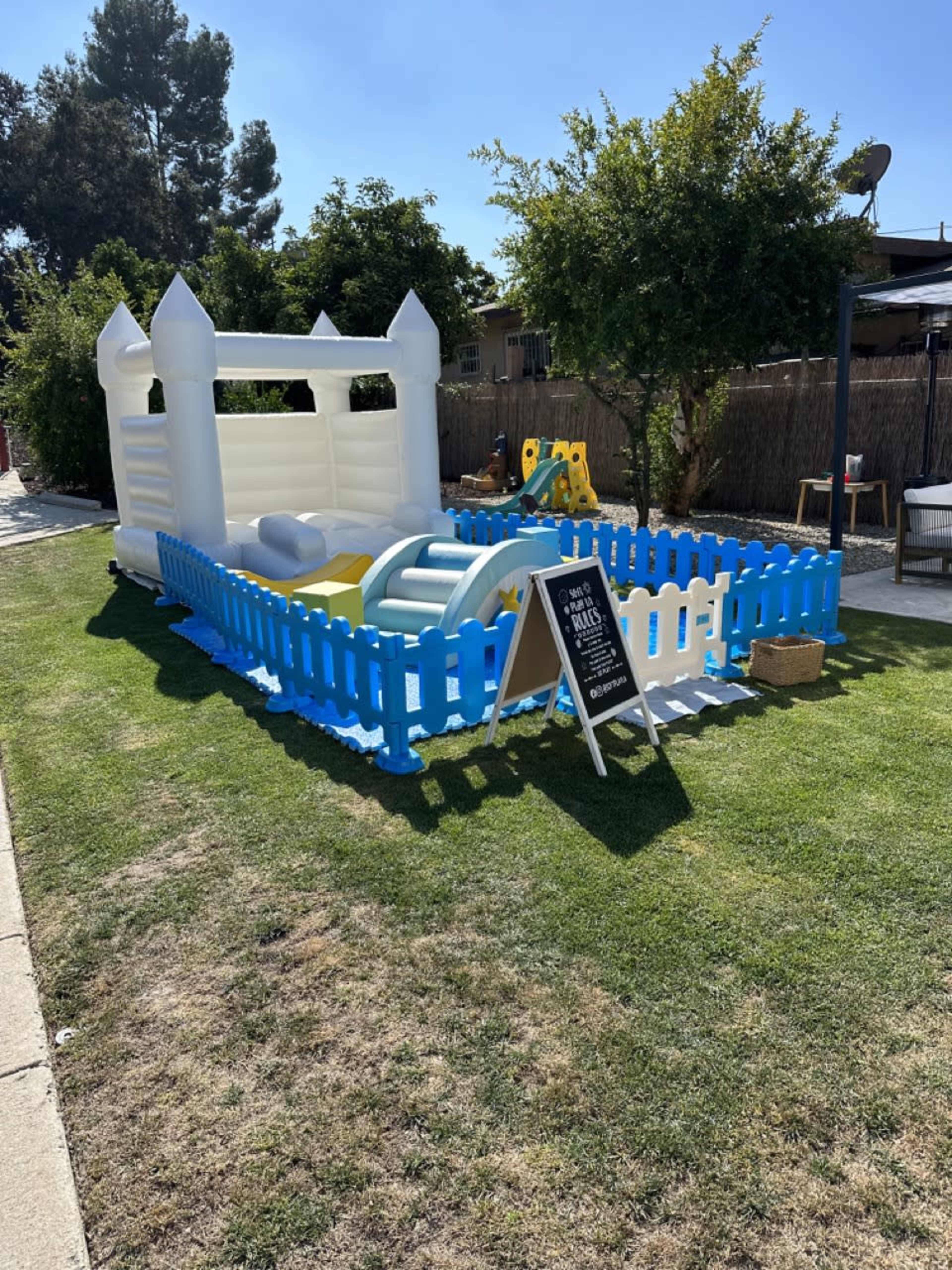 A colorful inflatable bounce house with a slide is enclosed by a blue and white fence in a grassy yard.
