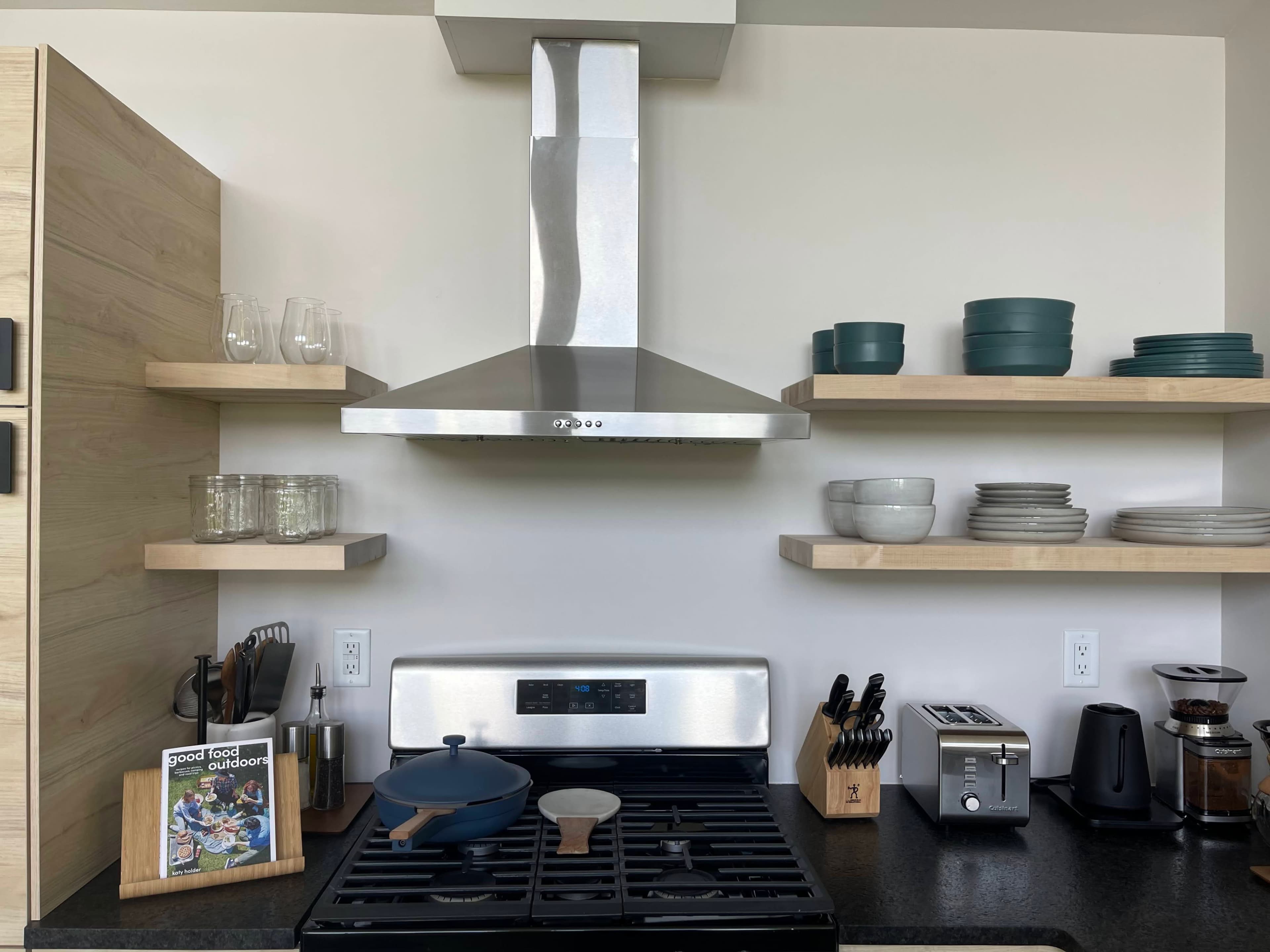 A modern kitchen with a stainless steel range hood, gas stove, shelves displaying various dishware, and organized kitchen utensils.