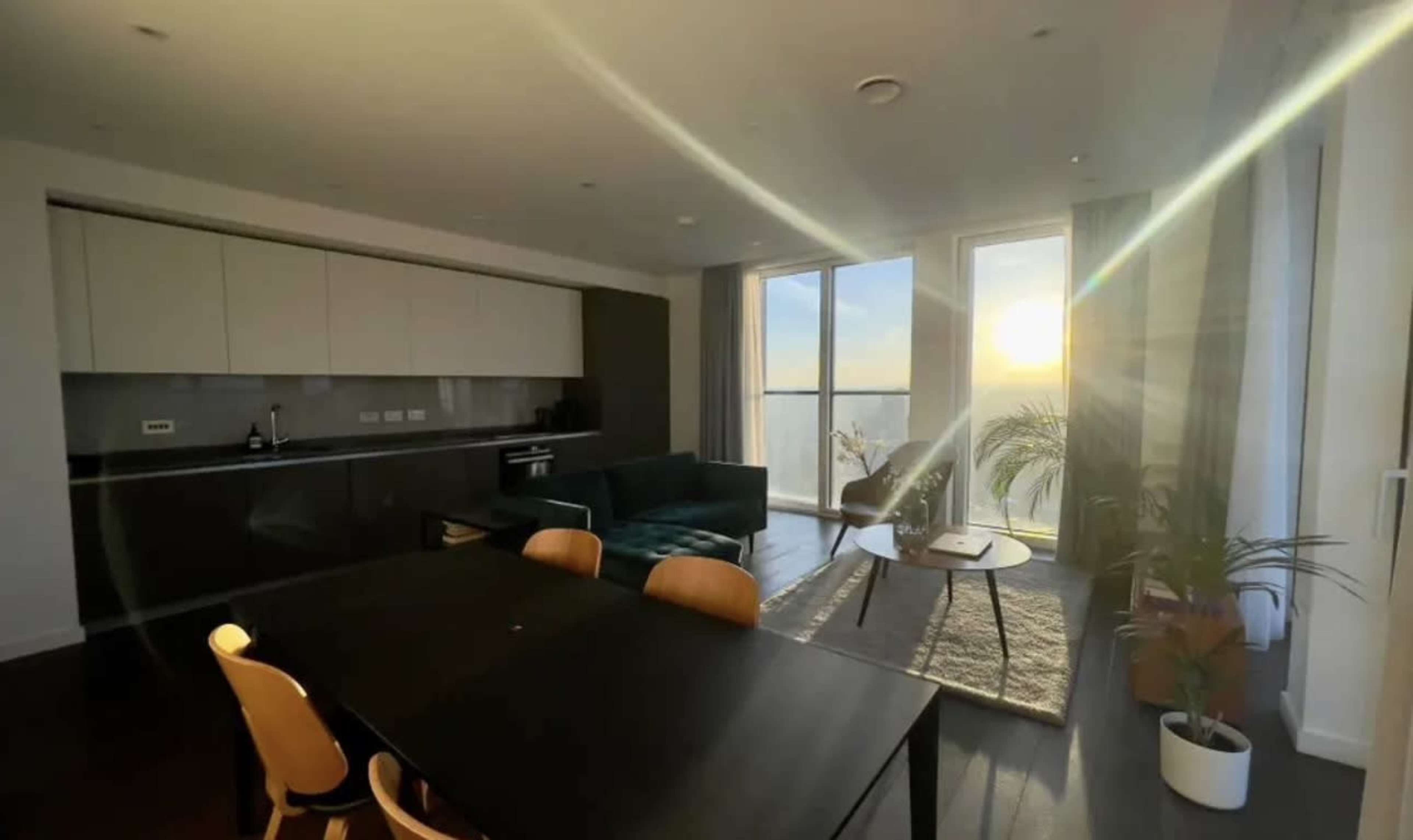 The image shows a modern kitchen and living area with a large window revealing a sunset, a dining table, and a green couch.