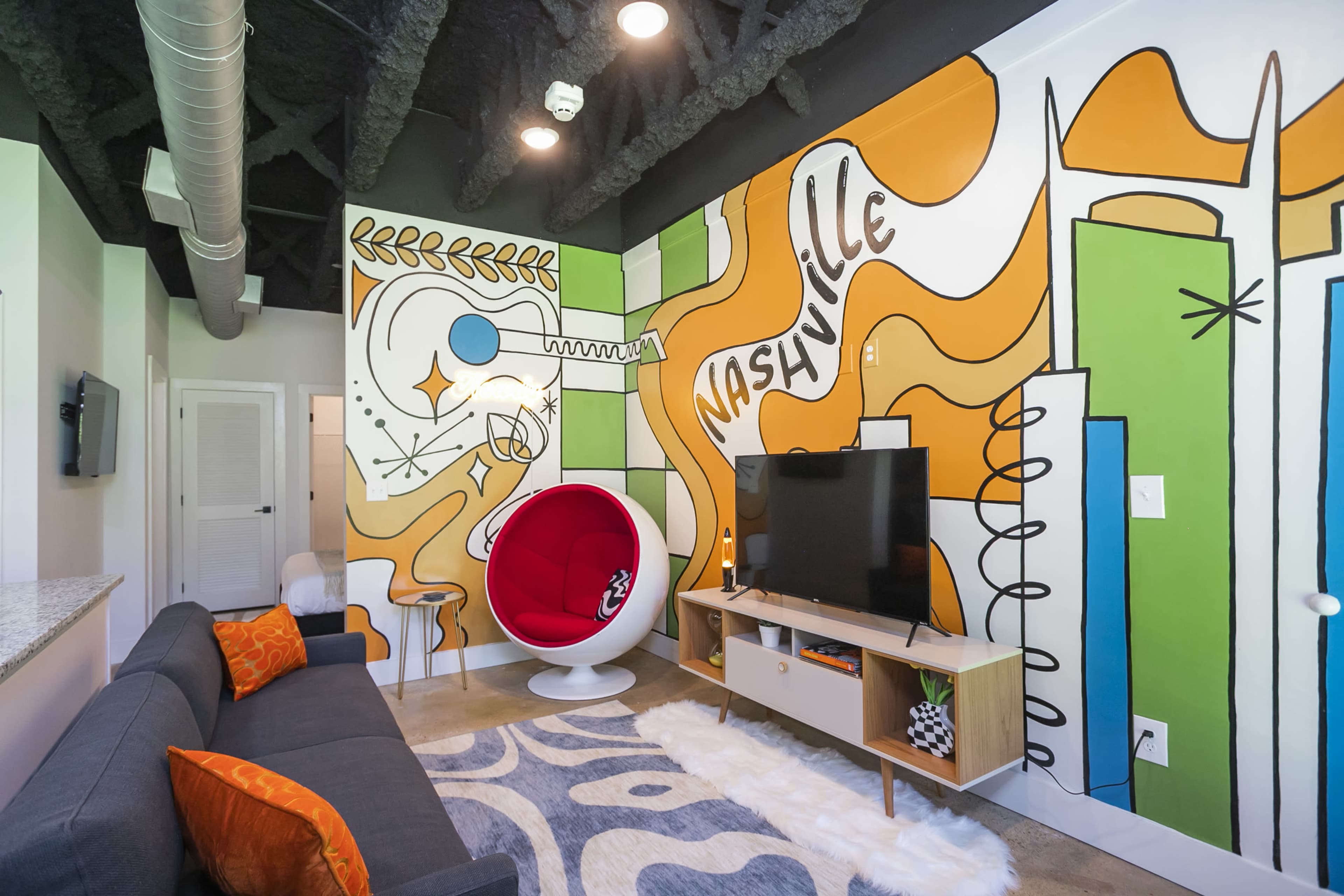 The room features a colorful mural on the walls, a modern white chair, a television stand, and a sofa with orange accents.