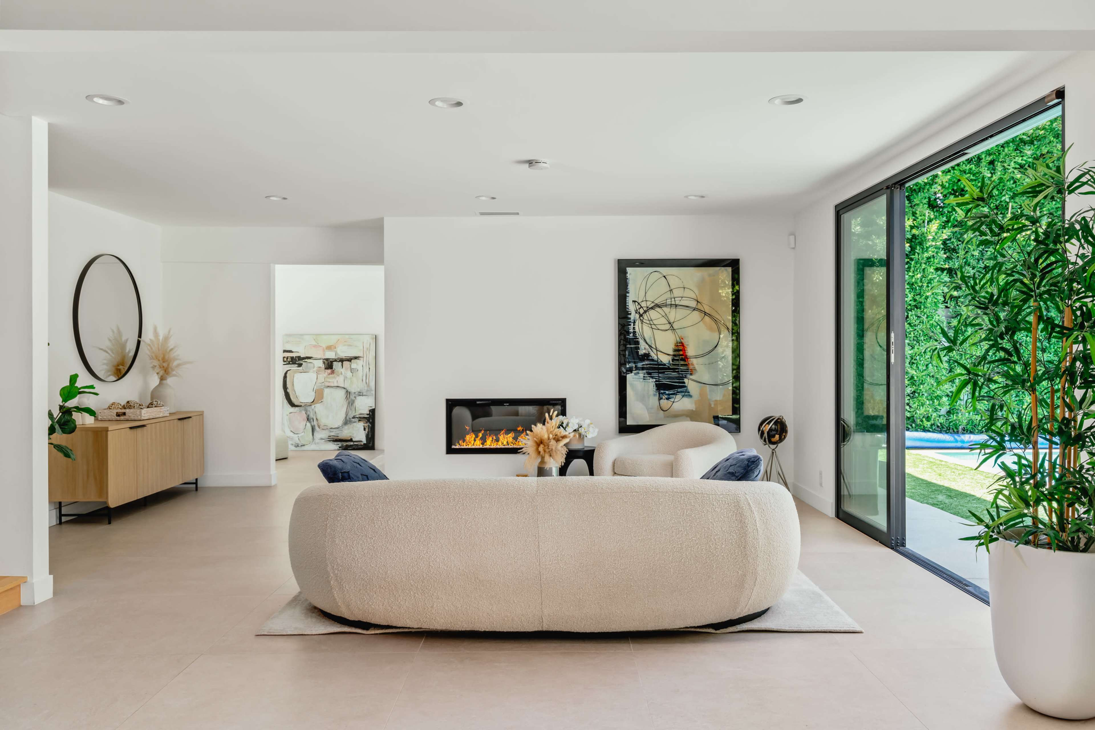 The living area features a large circular sofa, a minimalist sideboard, and glass doors leading to an outdoor space.